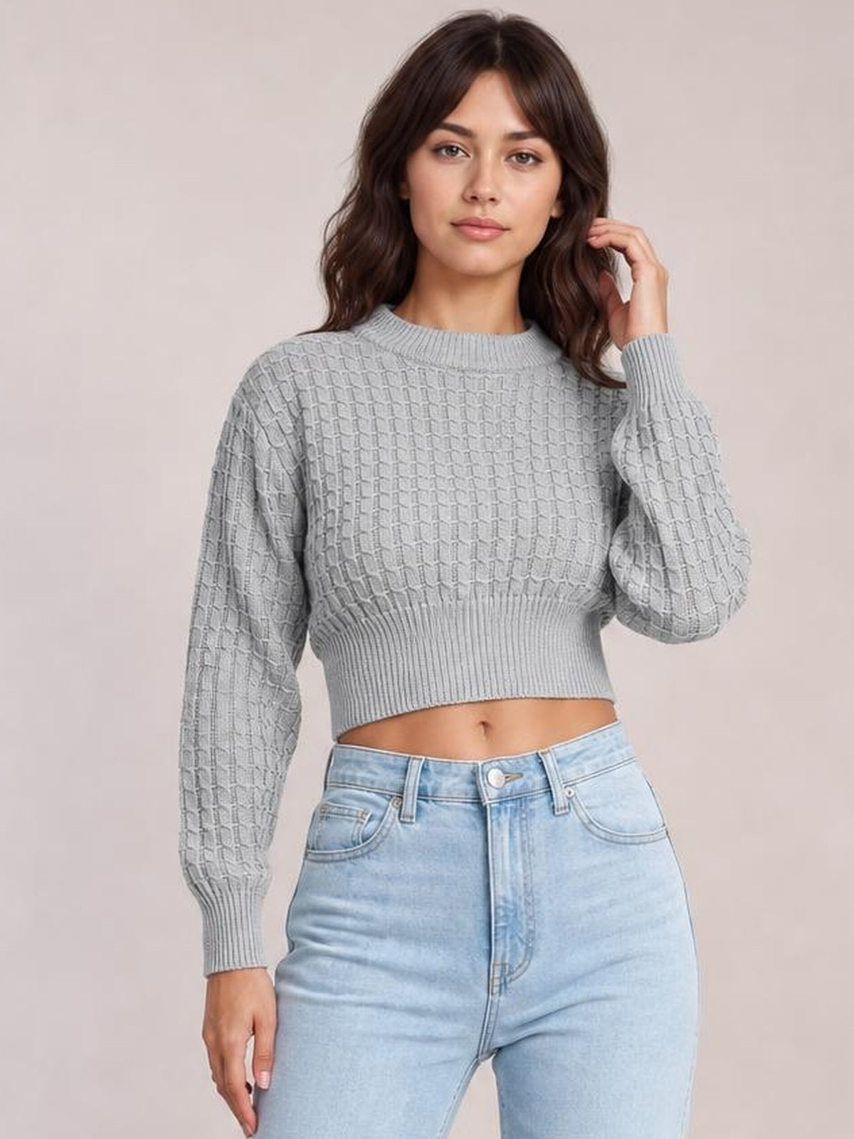 Model wears a light gray round neck long sleeve cropped sweater with textured knit construction, ribbed cuffs and hem, posing with one hand in her hair.