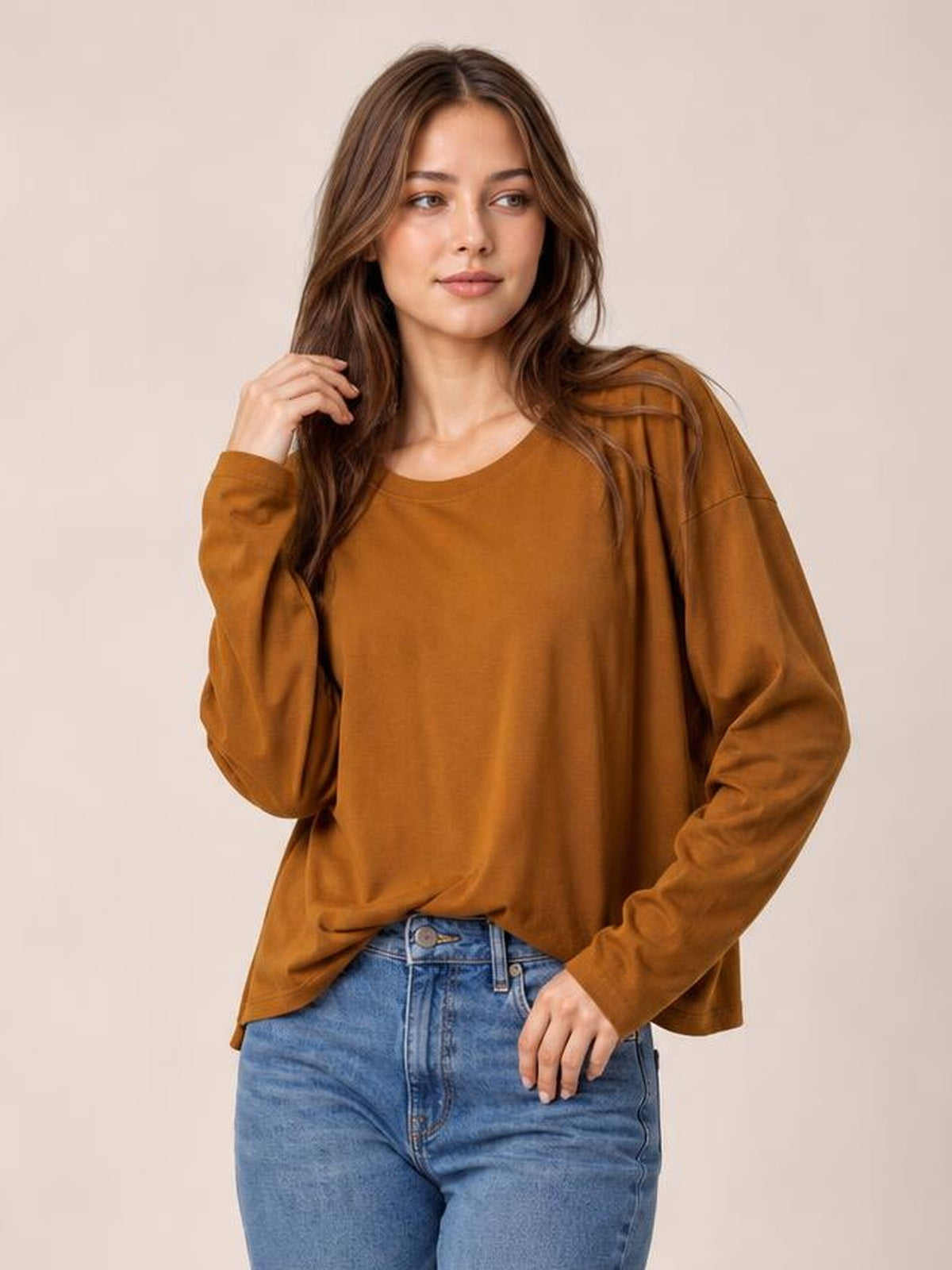 Model poses in a rust-brown round-neck long-sleeve cropped T-shirt with a relaxed silhouette and simple construction, one hand touching her hair.