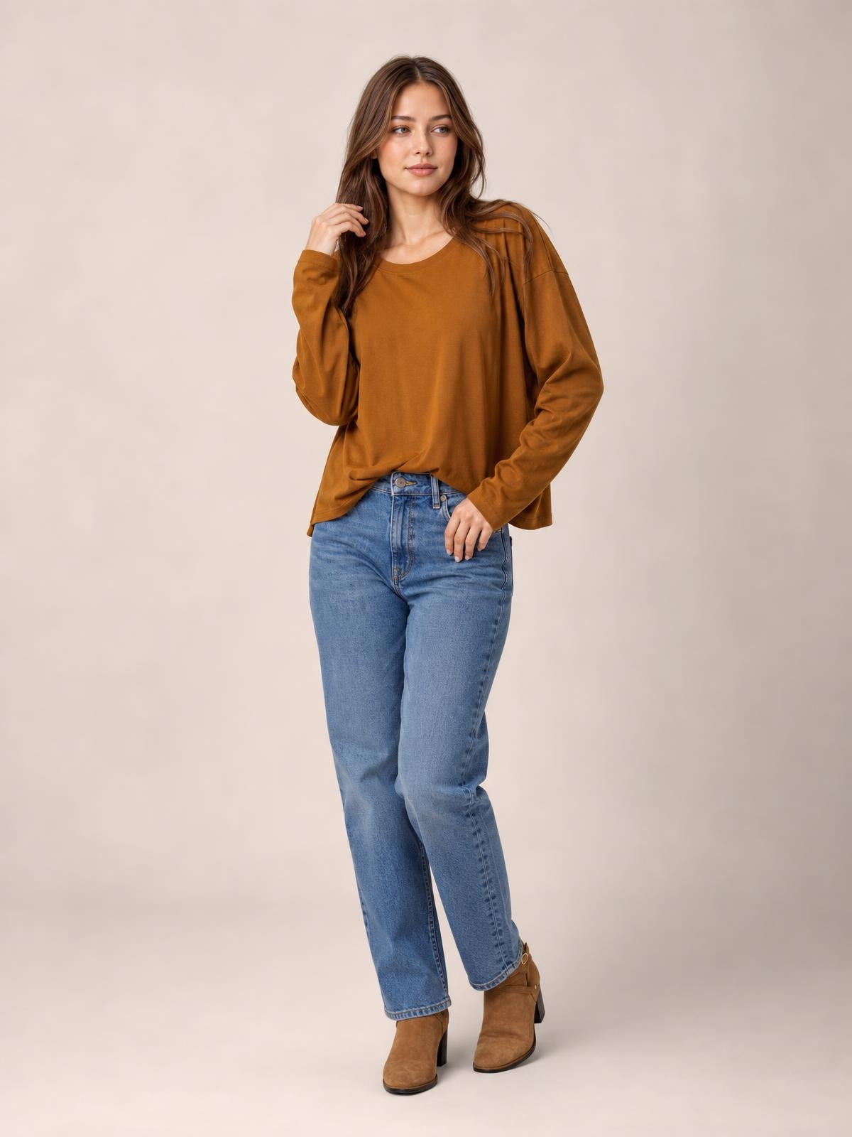 A model wears a brown round neck long sleeve cropped T-shirt with a relaxed silhouette, posing with one hand touching her hair.
