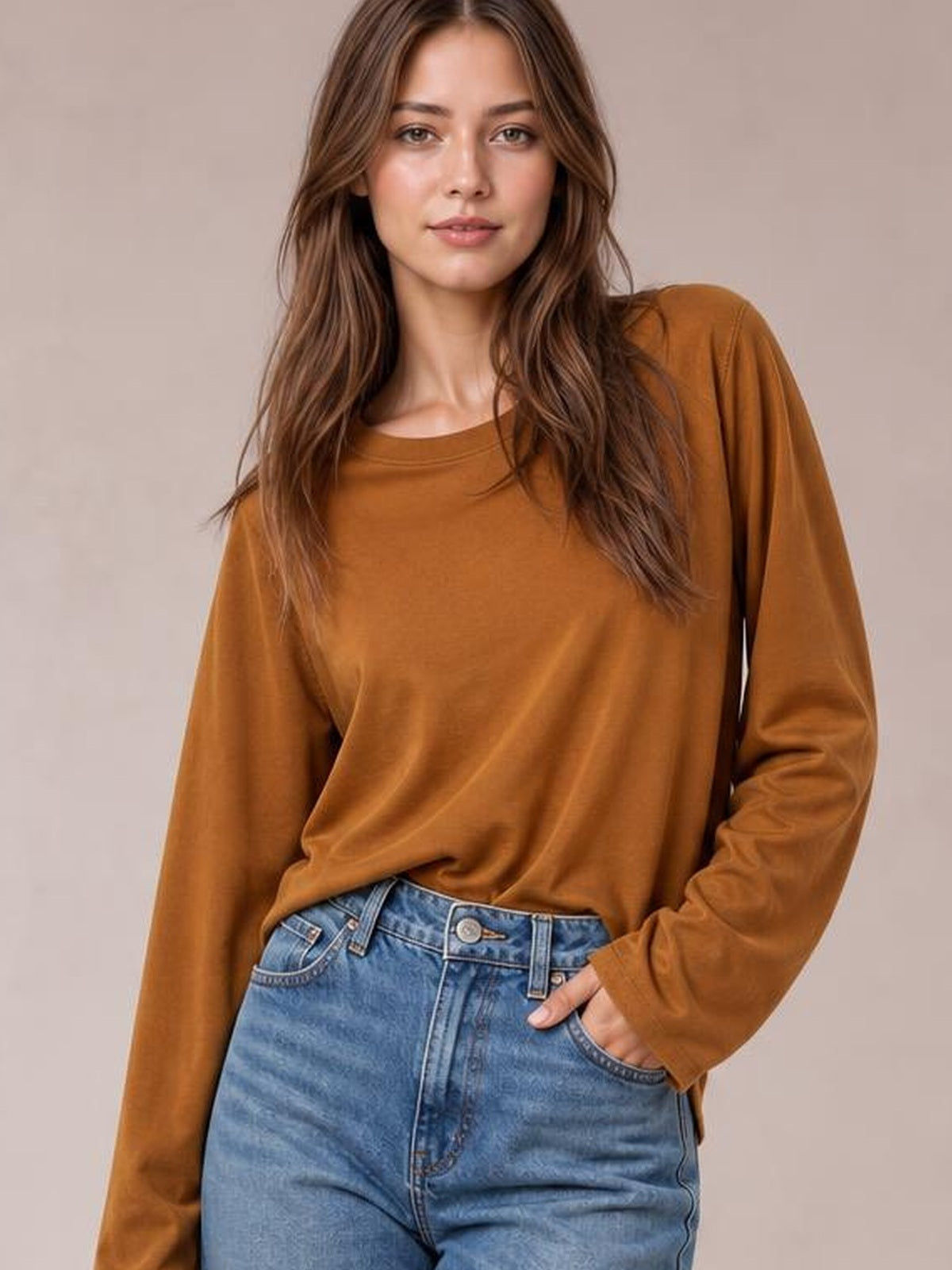 Model wearing a rust-brown round neck long sleeve cropped T-shirt with a relaxed silhouette, soft drape, and simple construction, posing with one hand in her jeans pocket.