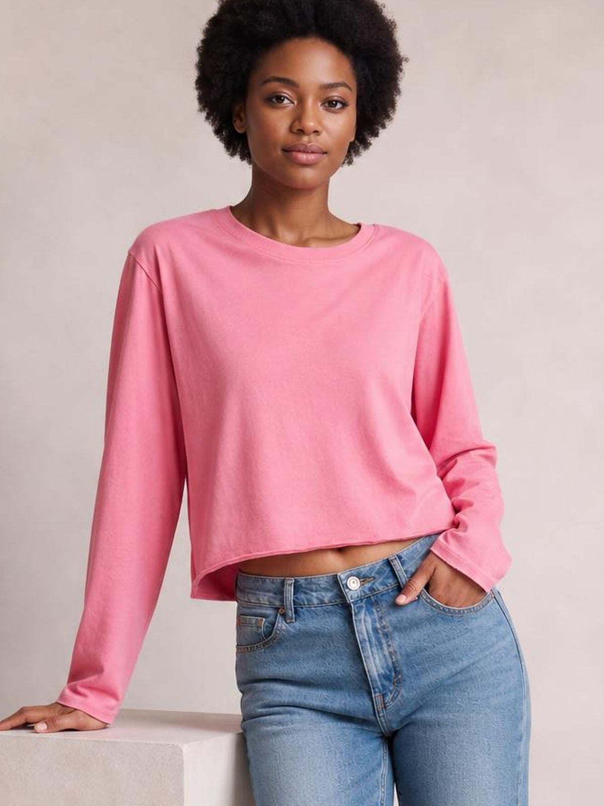 Model wearing a pink round-neck long-sleeve cropped T-shirt with a relaxed silhouette, raw hem, and one hand resting in her jeans pocket.