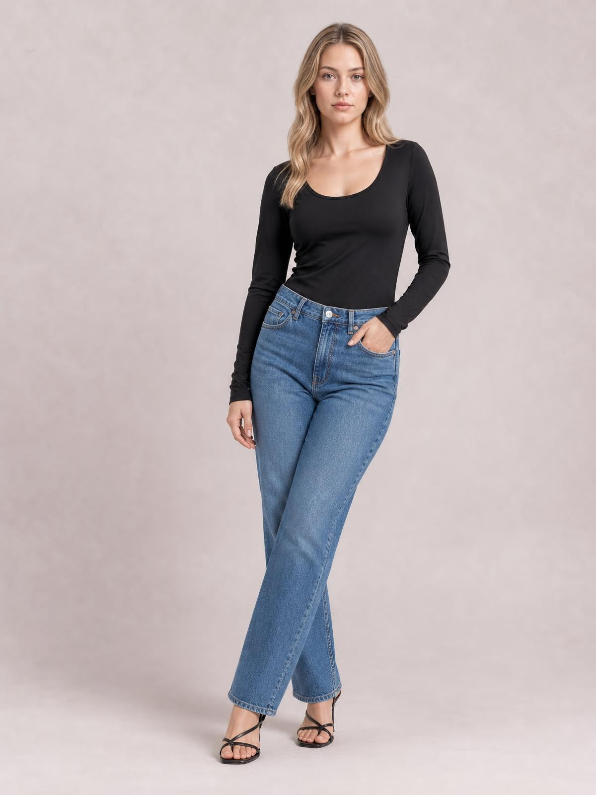 A model poses in a black round neck long sleeve T-shirt with a fitted silhouette, slightly stretchy opaque fabric, and one hand tucked into her jeans pocket.