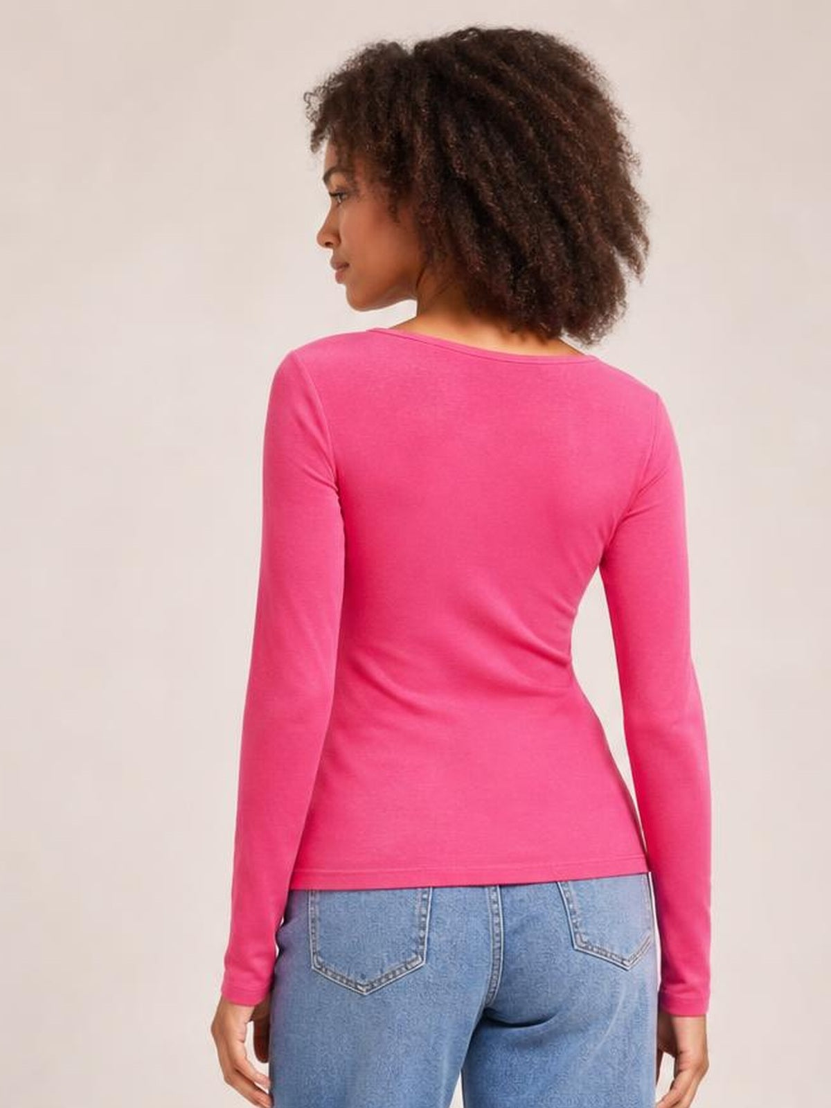 A model poses in a back view wearing a bright pink round-neck long-sleeve T-shirt with a smooth, fitted silhouette and hem-length construction.