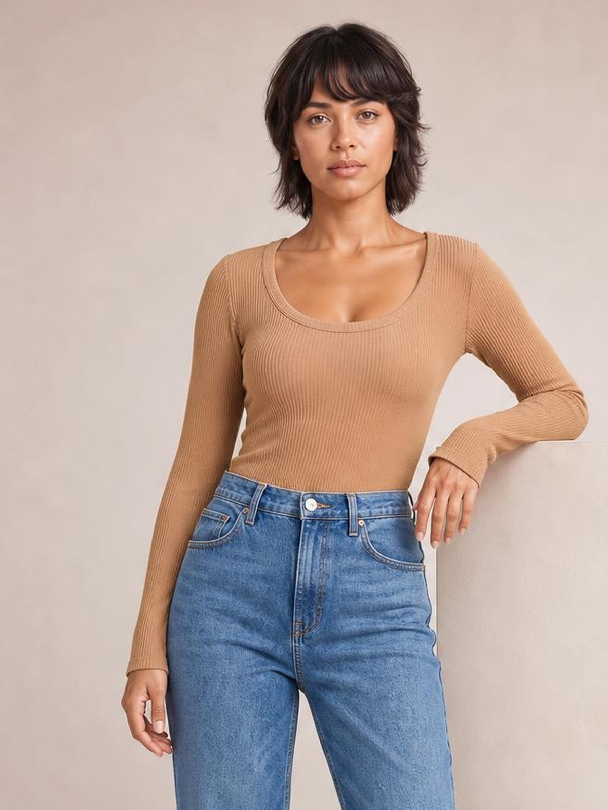 Model wearing a tan round-neck long-sleeve T-shirt with a fitted silhouette and fine ribbed construction, posing with one hand resting at her hip.