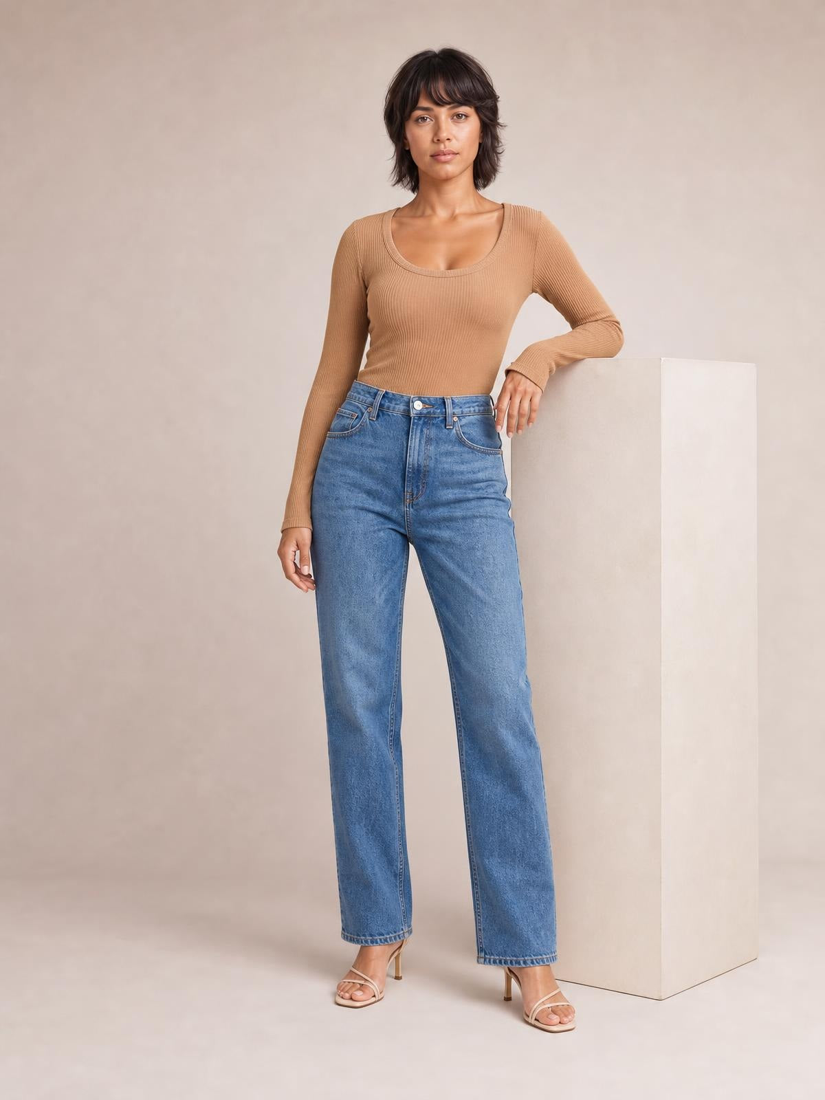 A model wears a tan round-neck long-sleeve T-shirt with a fitted, subtly ribbed silhouette, standing with one hand resting on a display block.
