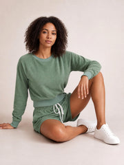 A seated model wears a sage green round neck long sleeve top with ribbed cuffs and hem, paired with drawstring shorts in a relaxed lounge set.