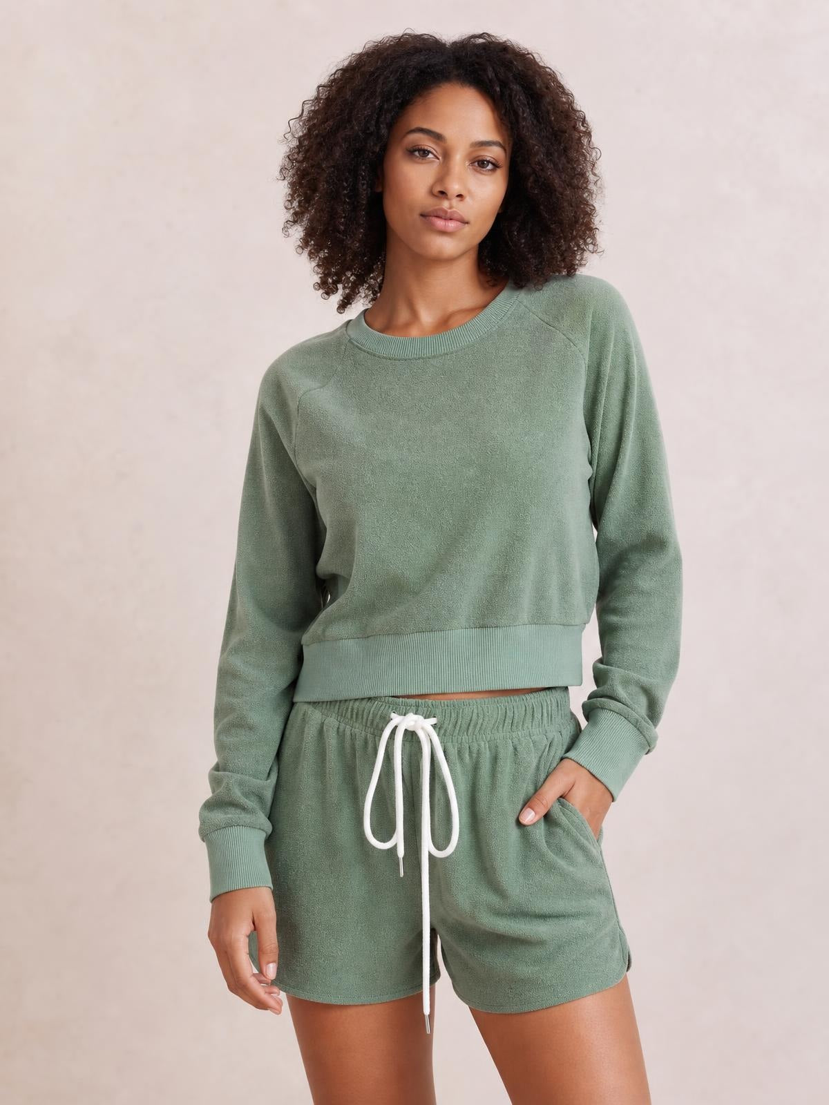Model poses in a sage green lounge set with a round-neck long-sleeve top, ribbed cuffs and hem, and drawstring shorts with pockets.