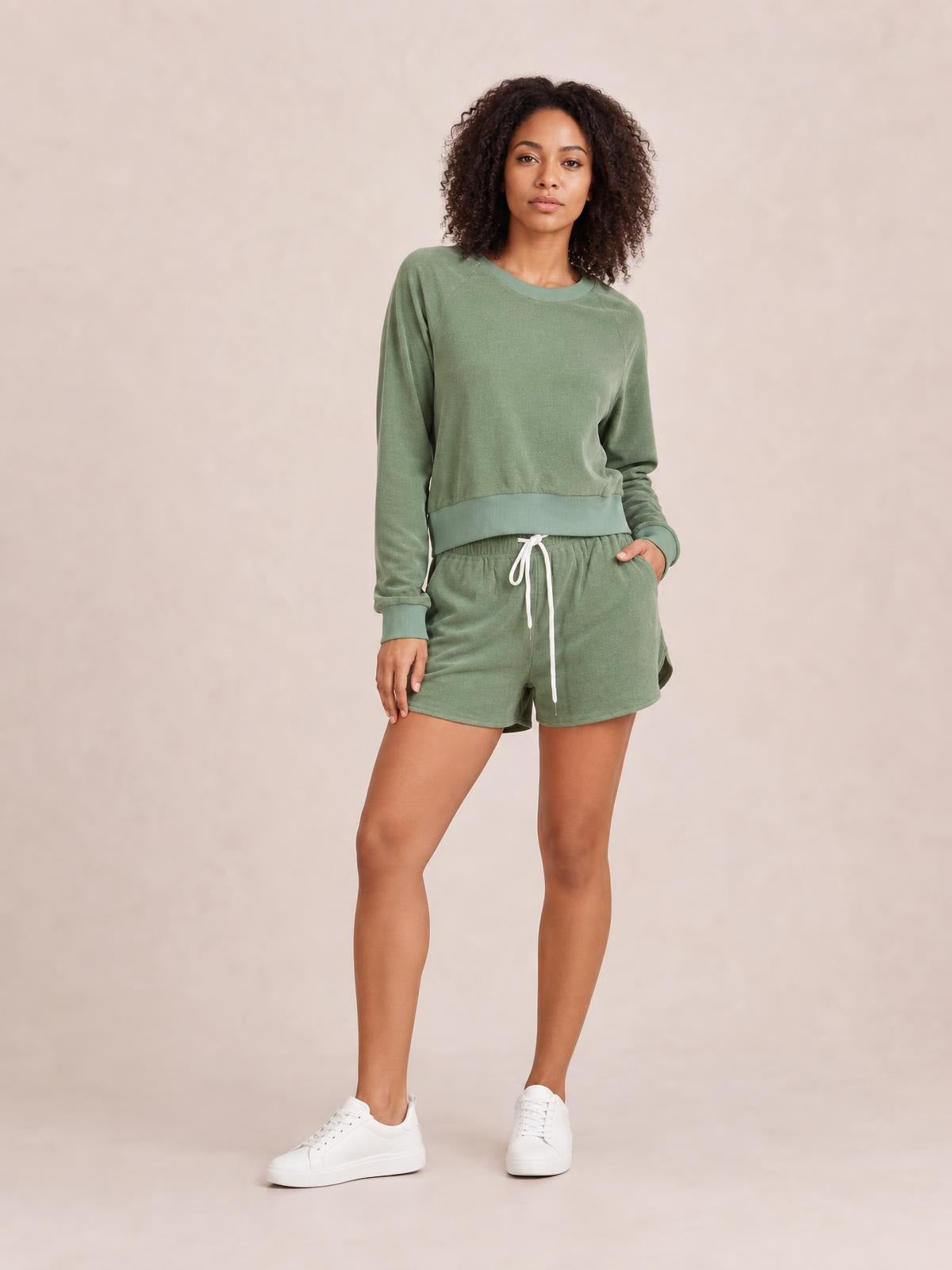 Model poses in a sage green round neck long sleeve top and matching drawstring shorts lounge set with ribbed cuffs and relaxed silhouette.