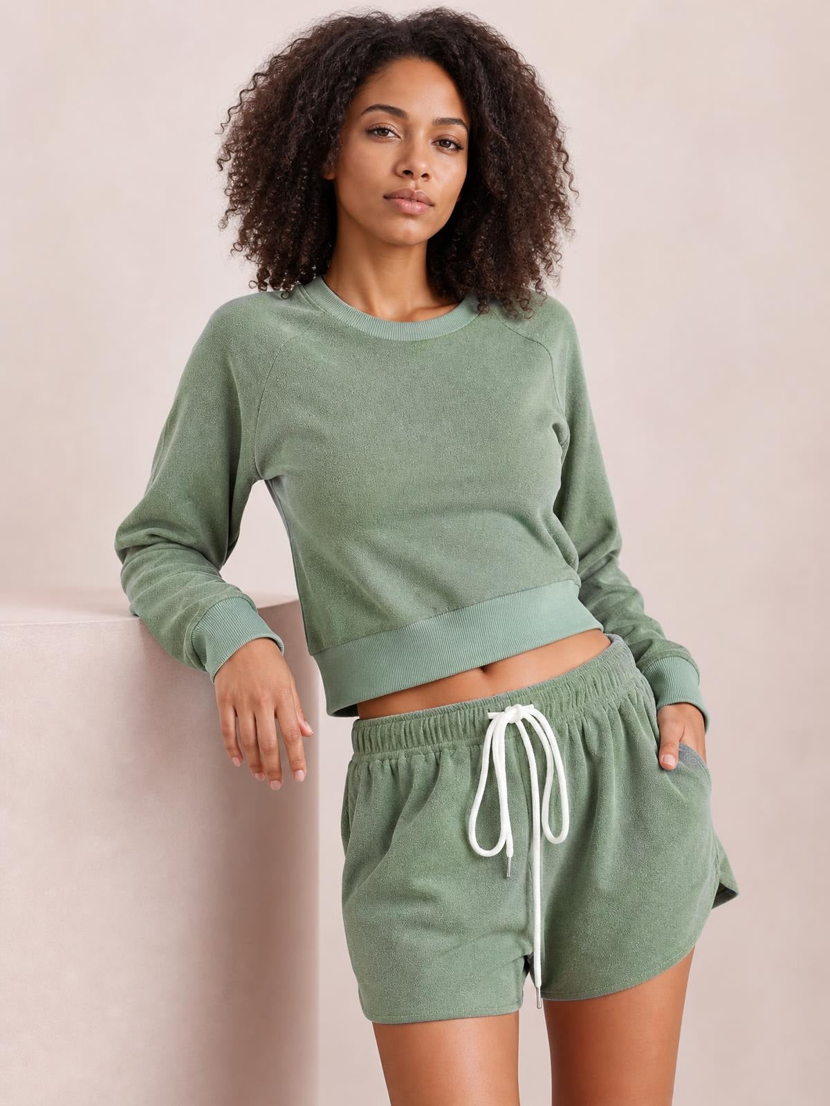 Model leans casually in a sage green lounge set with a round-neck long-sleeve top, ribbed cuffs and hem, and relaxed drawstring shorts.