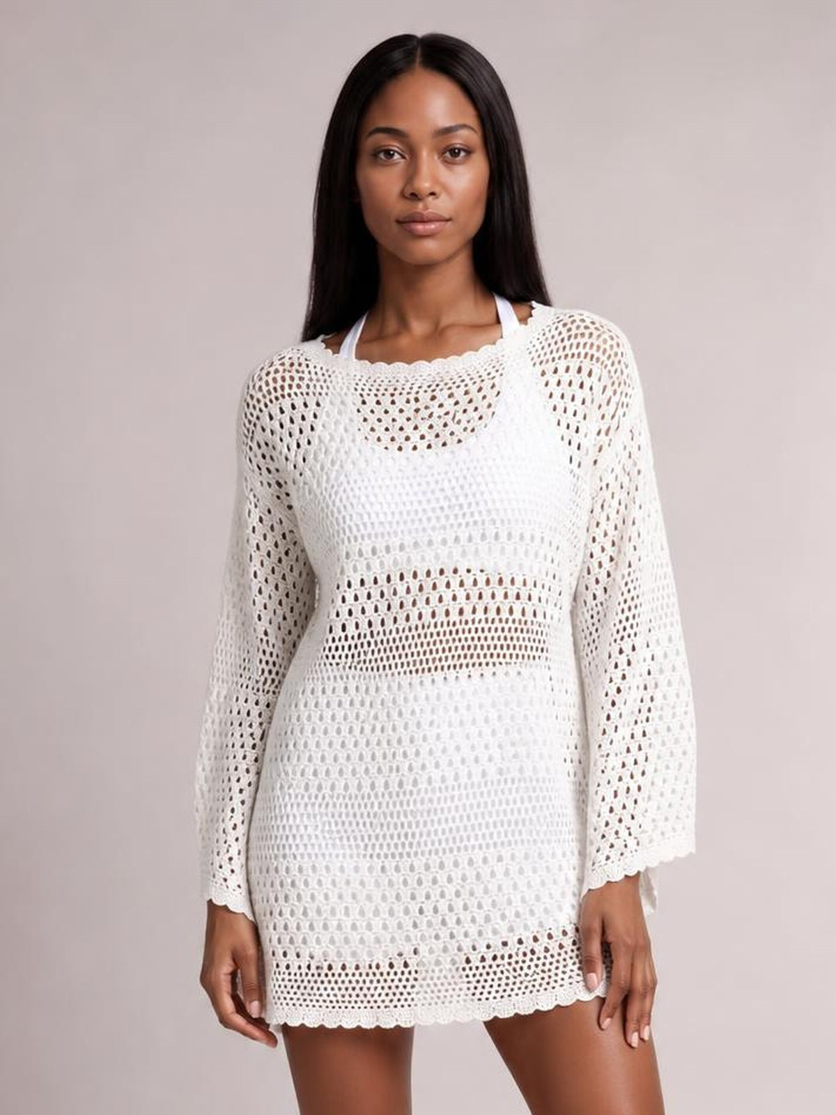 A model stands wearing a white round-neck openwork cover-up with cutout crochet-style texture, long sleeves, a relaxed tunic silhouette, and scalloped edges.