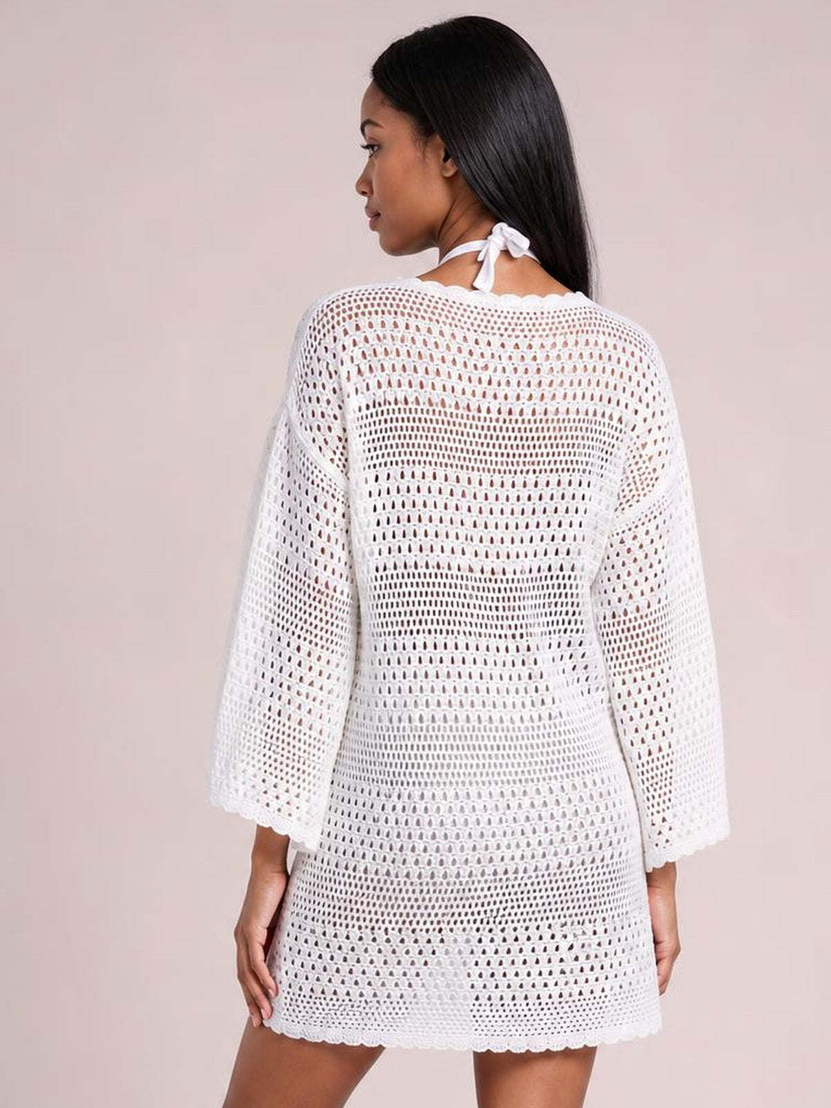 A model looks over her shoulder in a white round-neck openwork cover-up with long sleeves, a relaxed silhouette, cutout construction, and scalloped hem.