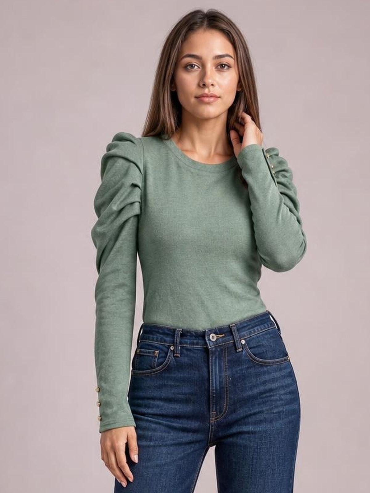 Model wearing a sage green round-neck blouse with voluminous puff sleeves, ruched construction, a fitted silhouette, and buttoned cuffs, posing with one hand near her hair.