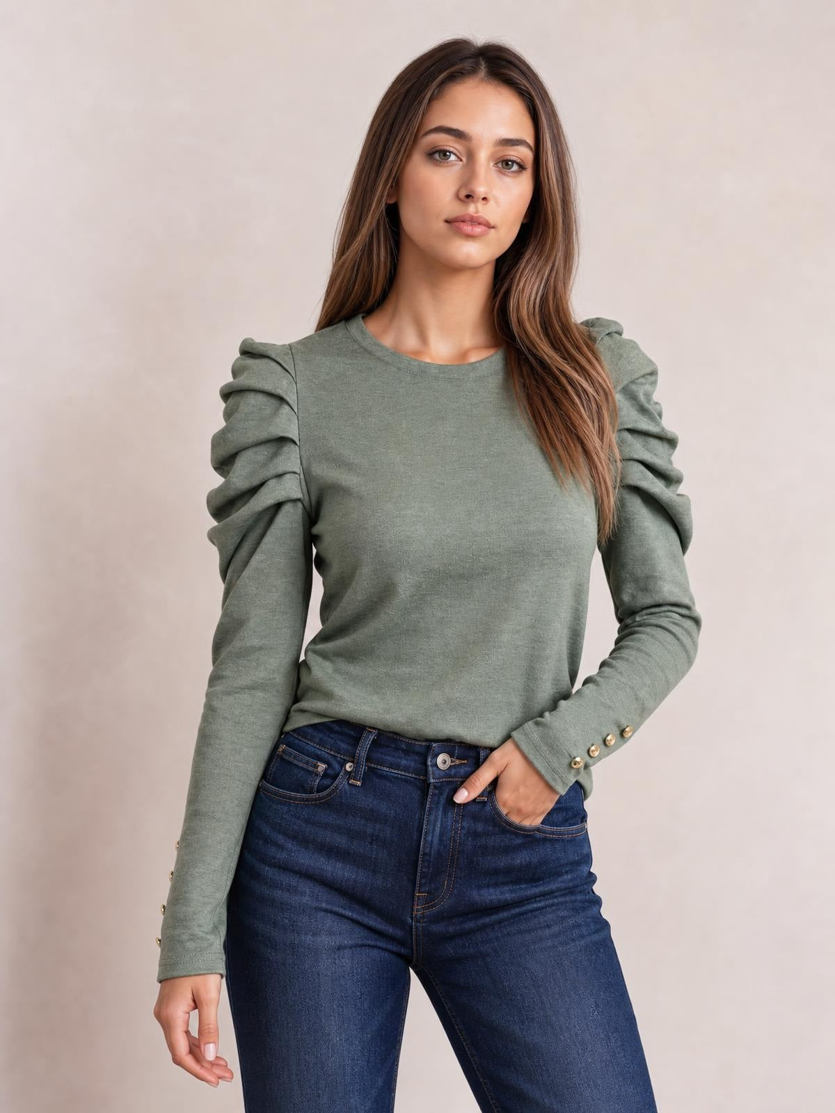 A model poses in a sage green round-neck blouse with gathered puff sleeves, a fitted silhouette, and buttoned cuffs, one hand resting in her jeans pocket.