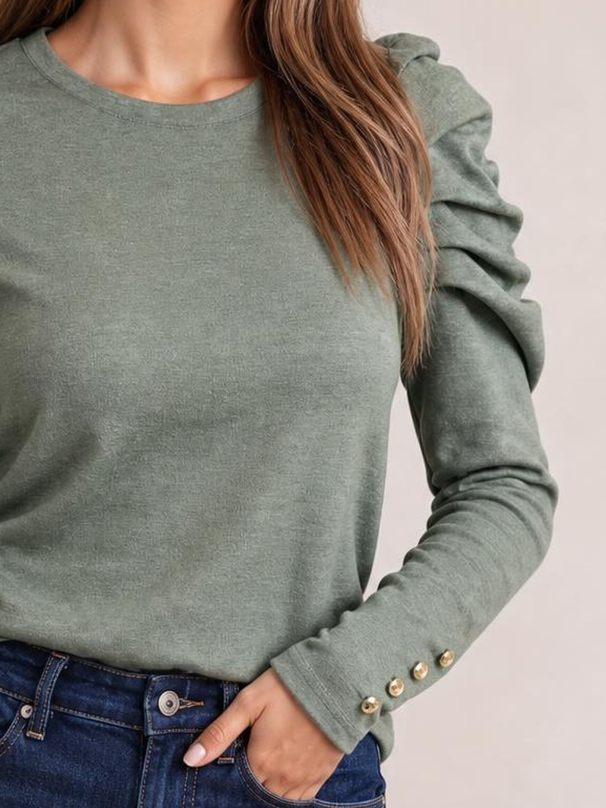 Model wearing a sage green round-neck blouse with voluminous puff sleeves, buttoned cuffs, and a relaxed silhouette, posing with one hand in her jeans pocket.