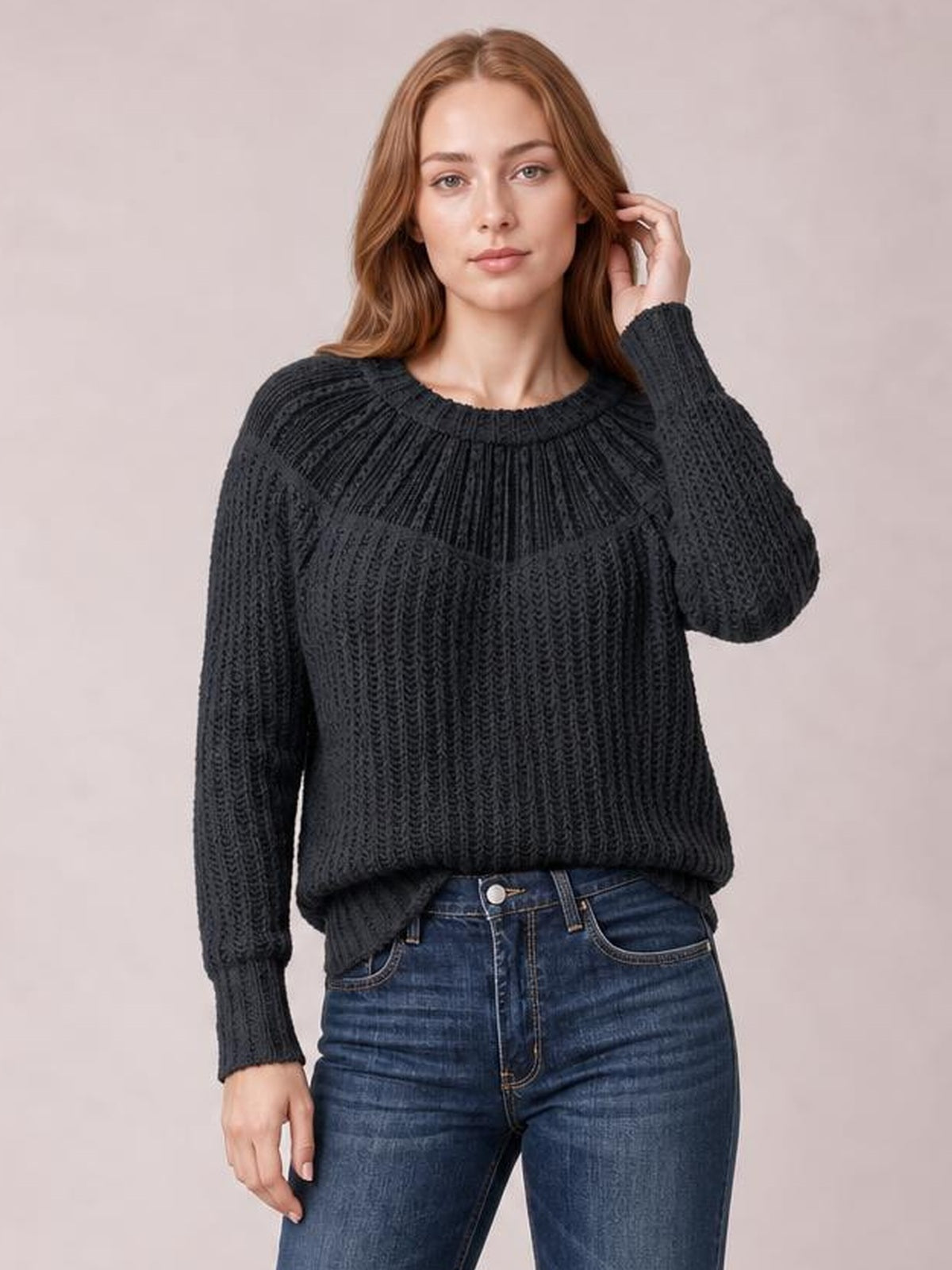 A model poses in a dark charcoal round-neck rib-knit sweater with textured vertical construction, long sleeves, and a relaxed silhouette while touching her hair.