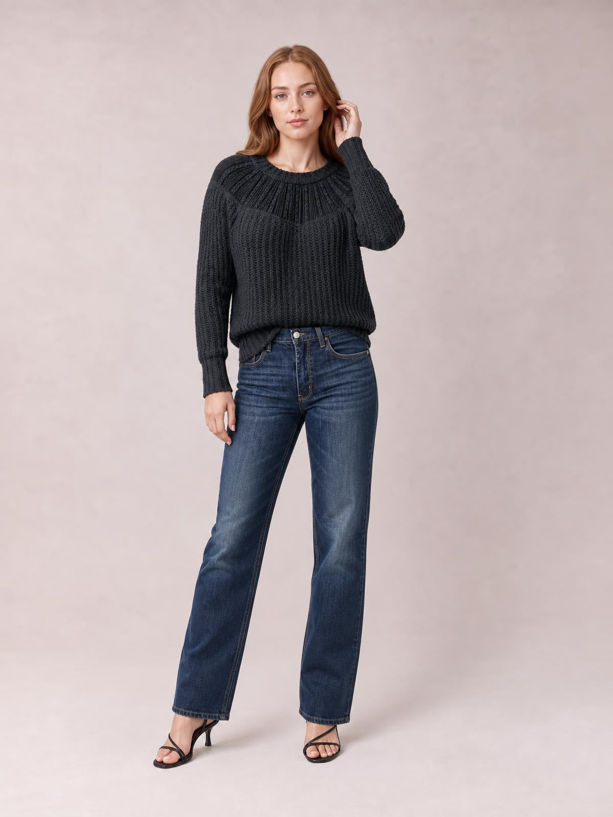 Model wears a black round-neck rib-knit sweater with long sleeves, textured vertical stitching, and a relaxed silhouette, posing with one hand near her hair.