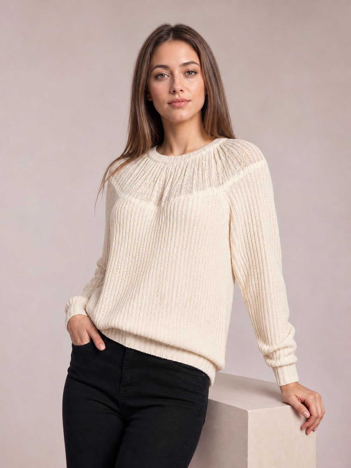 Model wearing an ivory round neck rib-knit sweater with a relaxed silhouette, long sleeves, ribbed cuffs and hem, leaning with one hand in a pocket.