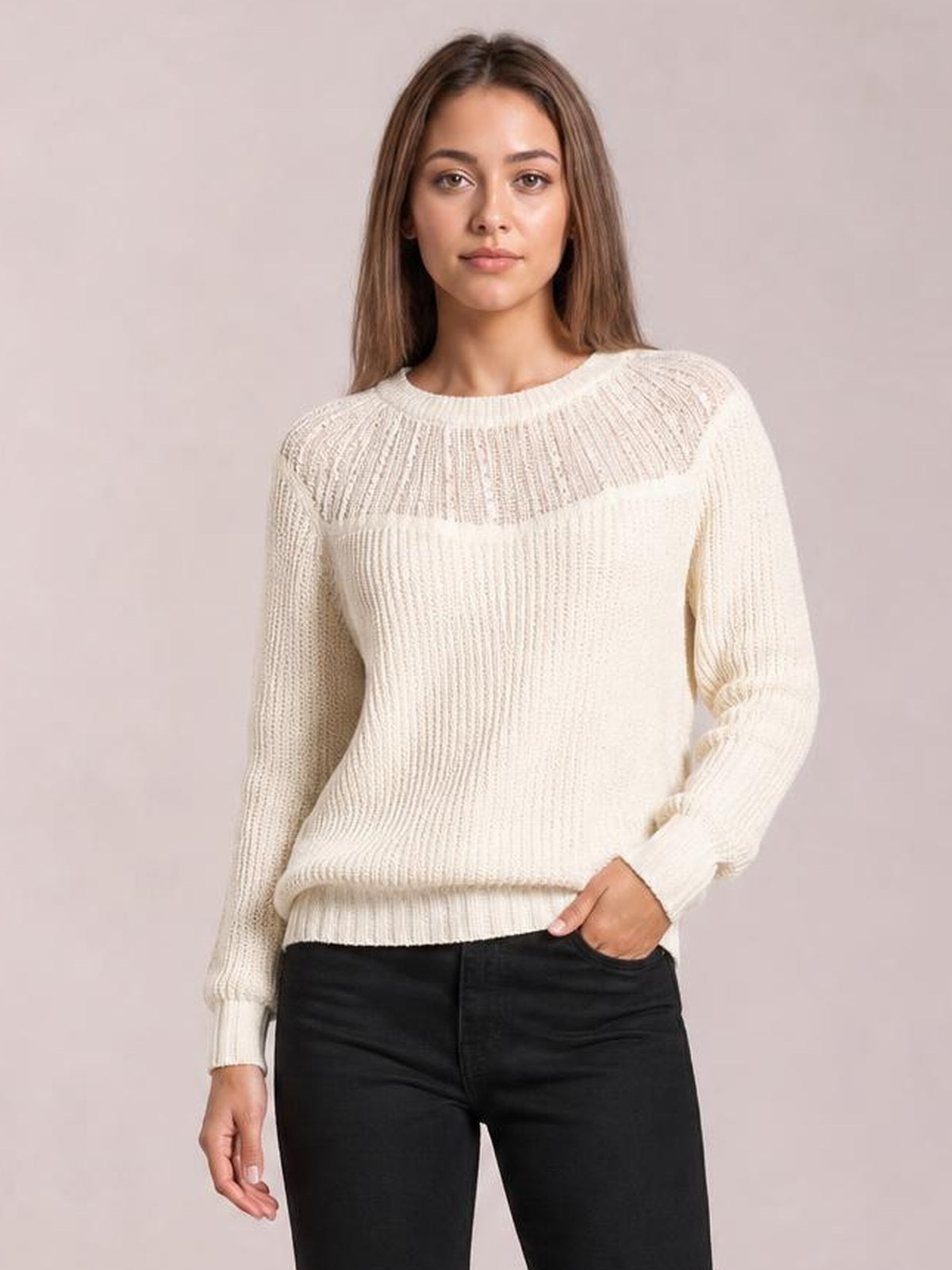A model stands with one hand in her pocket wearing a cream round-neck rib-knit sweater with long sleeves, a fitted silhouette, and semi-sheer open-knit texture.