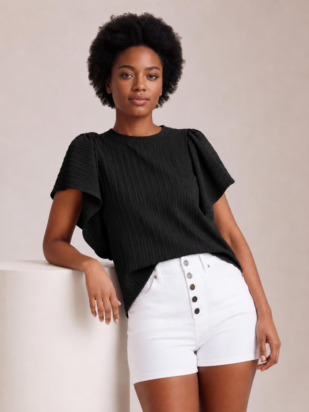 A model leans against a surface wearing a black round neck top with textured construction, short ruffle sleeves, and a relaxed, slightly loose silhouette.