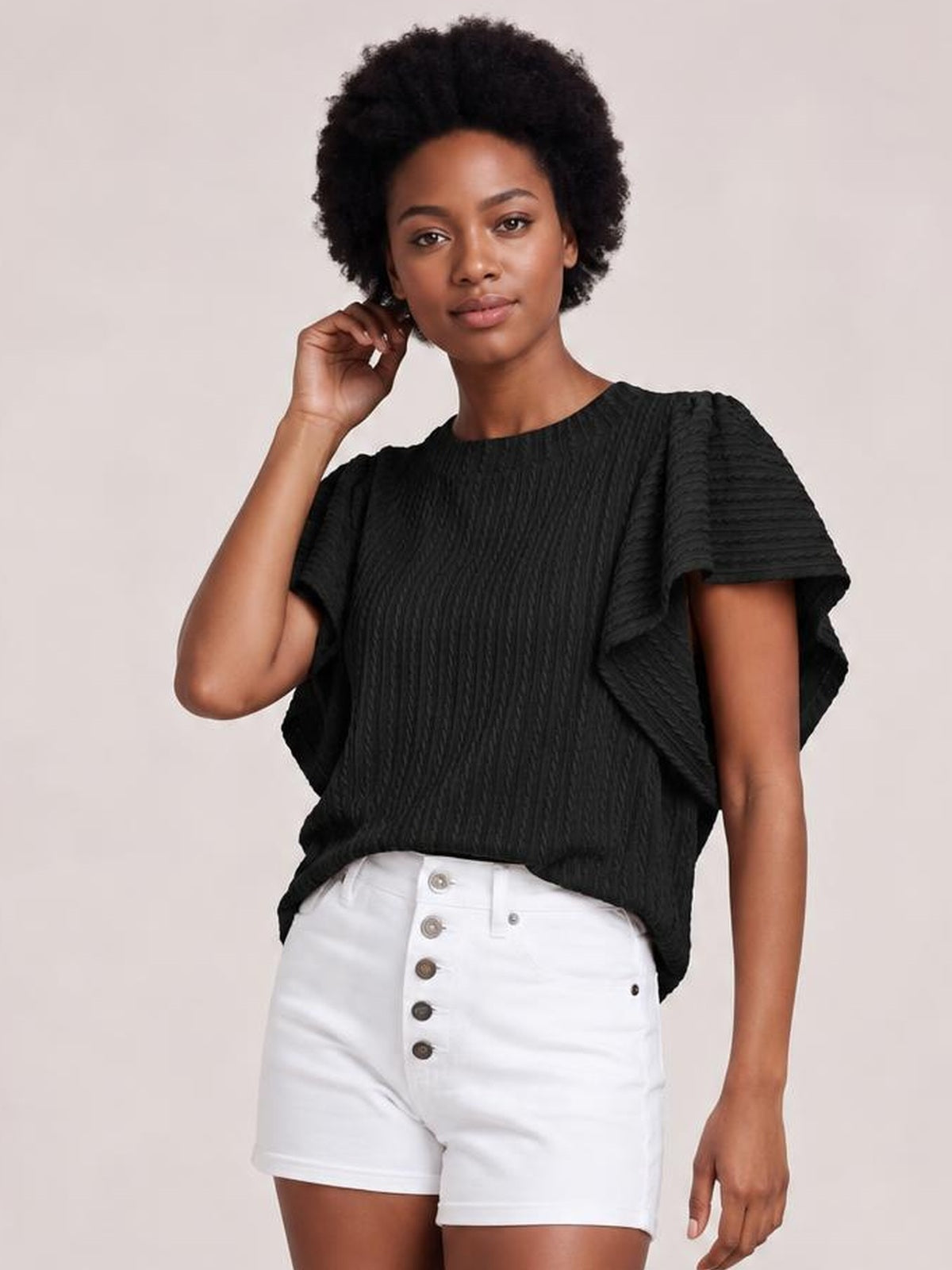 A model poses in a black round neck top with a relaxed silhouette, textured fabric, and layered ruffle sleeves, touching her hair with one hand.
