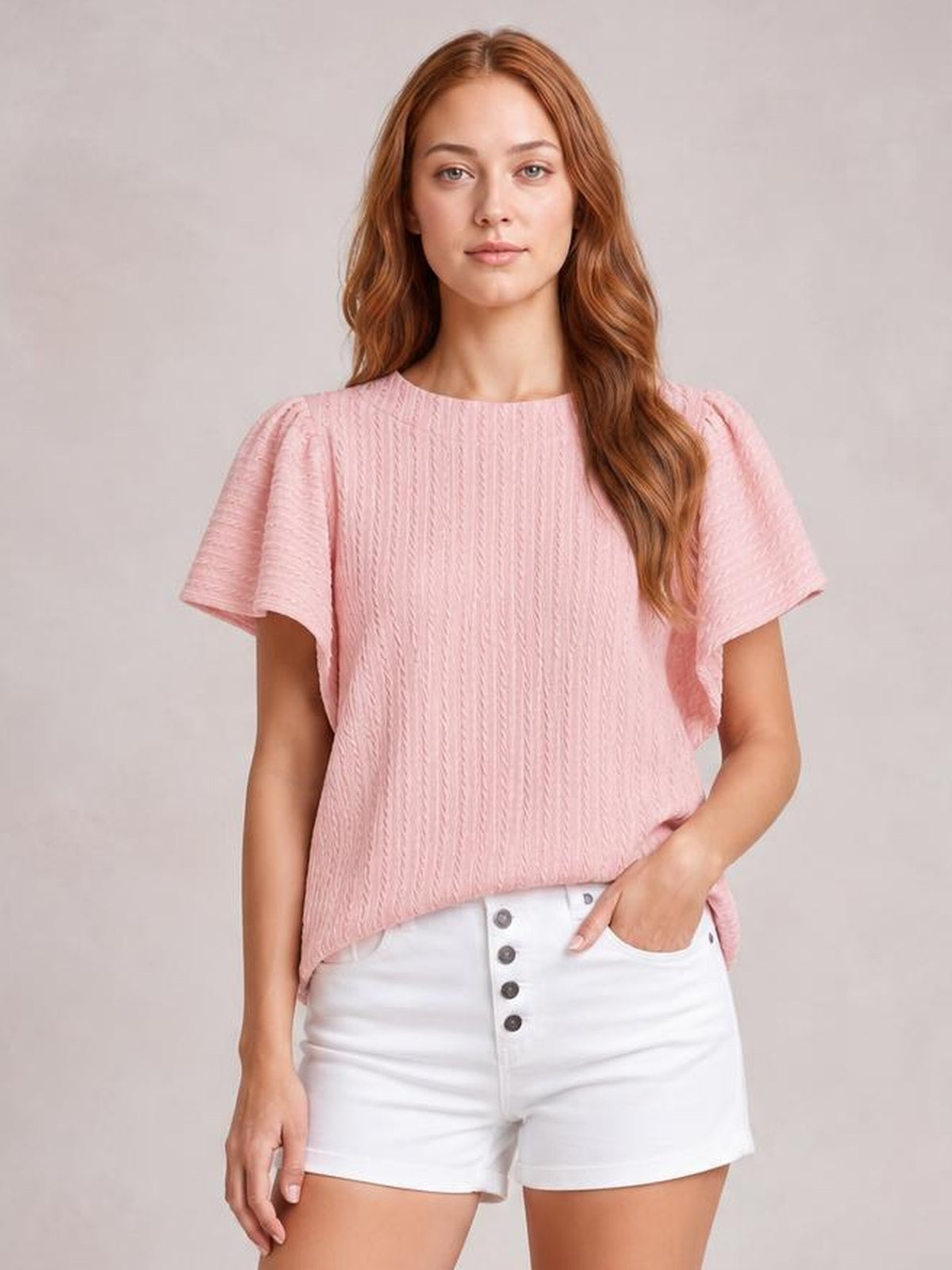 A model wears a pink round neck ruffle sleeve top with a textured vertical pattern, relaxed straight silhouette, and one hand resting in her shorts pocket.