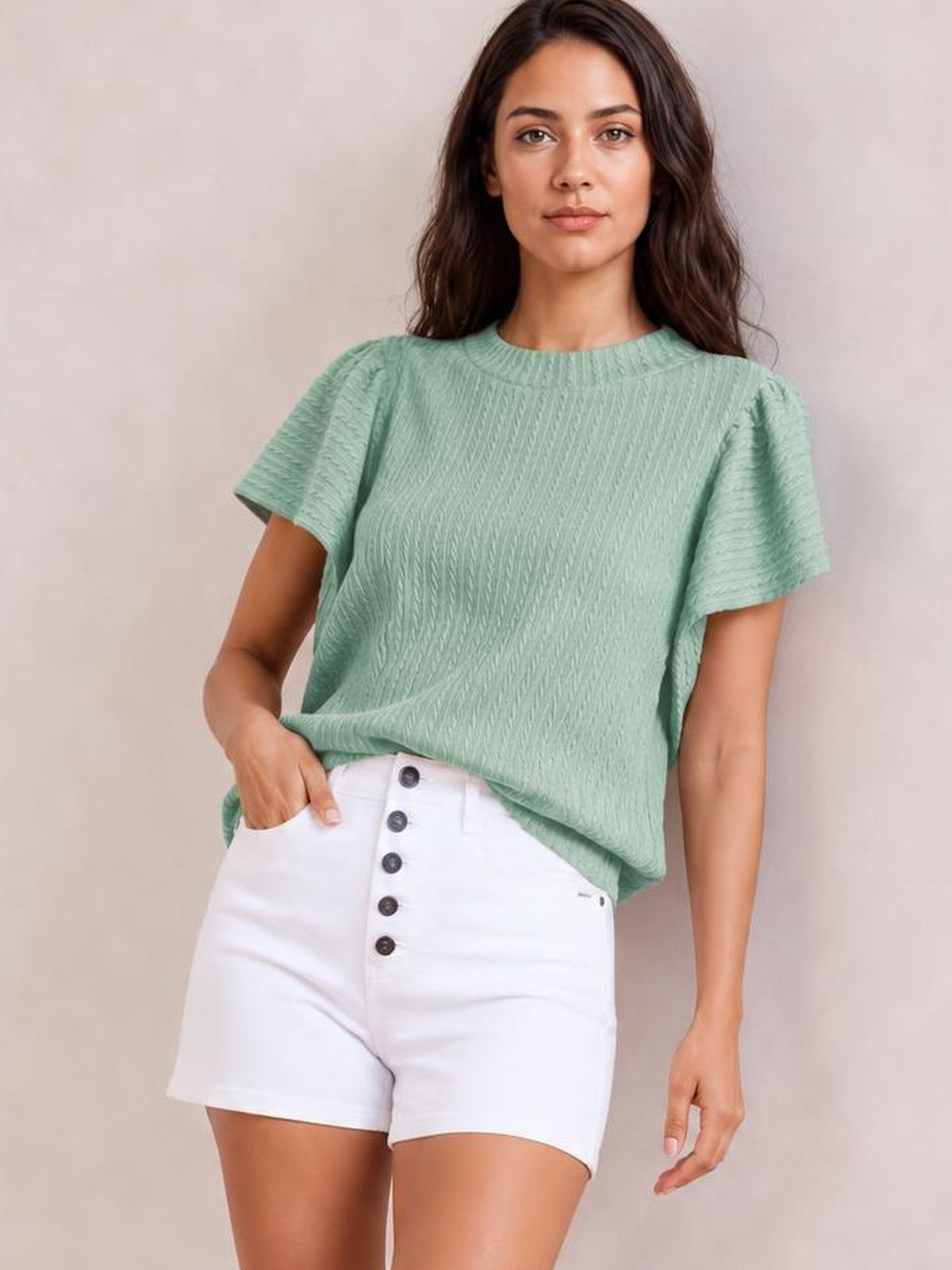 A model wears a mint green round-neck top with softly ruffled short sleeves, a textured finish, relaxed silhouette, and slight stretch, posing with one hand in her shorts pocket.