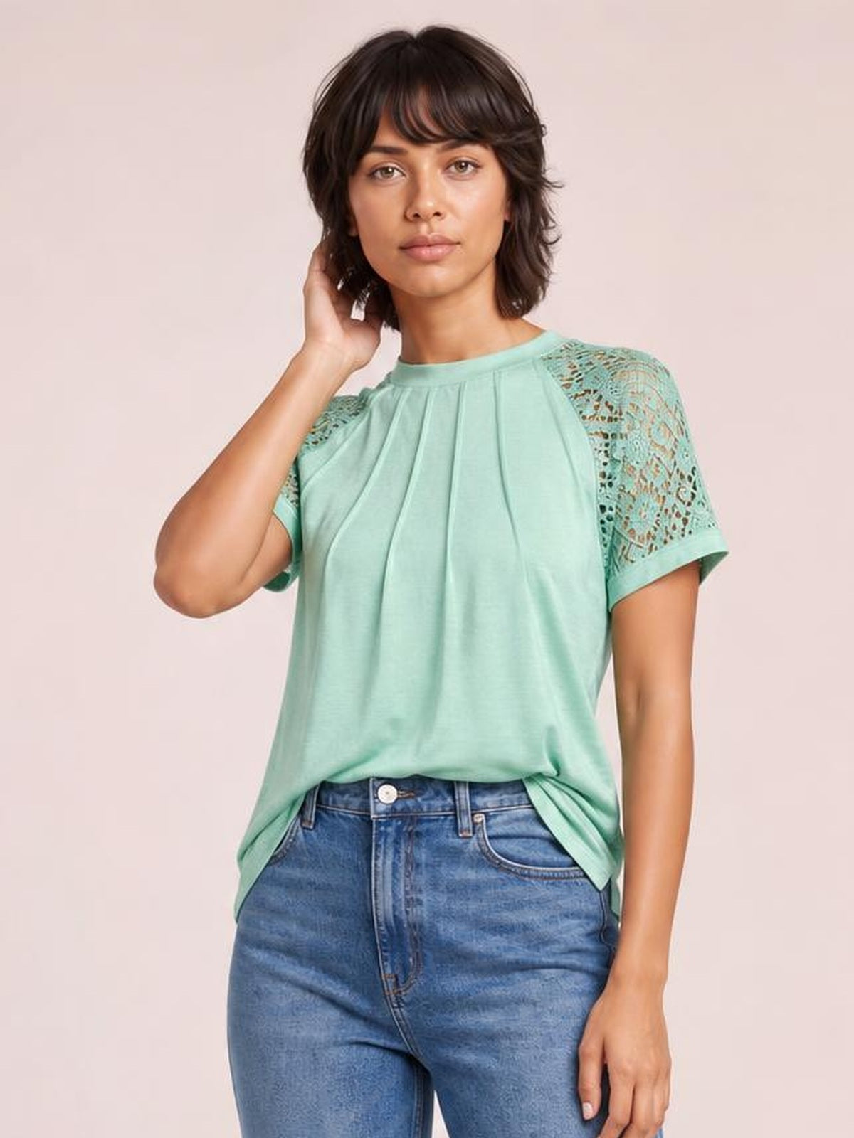 Model wears a mint green round neck short sleeve T-shirt with pleated front, lace sleeve panels, and a relaxed silhouette, posing with one hand in her hair.