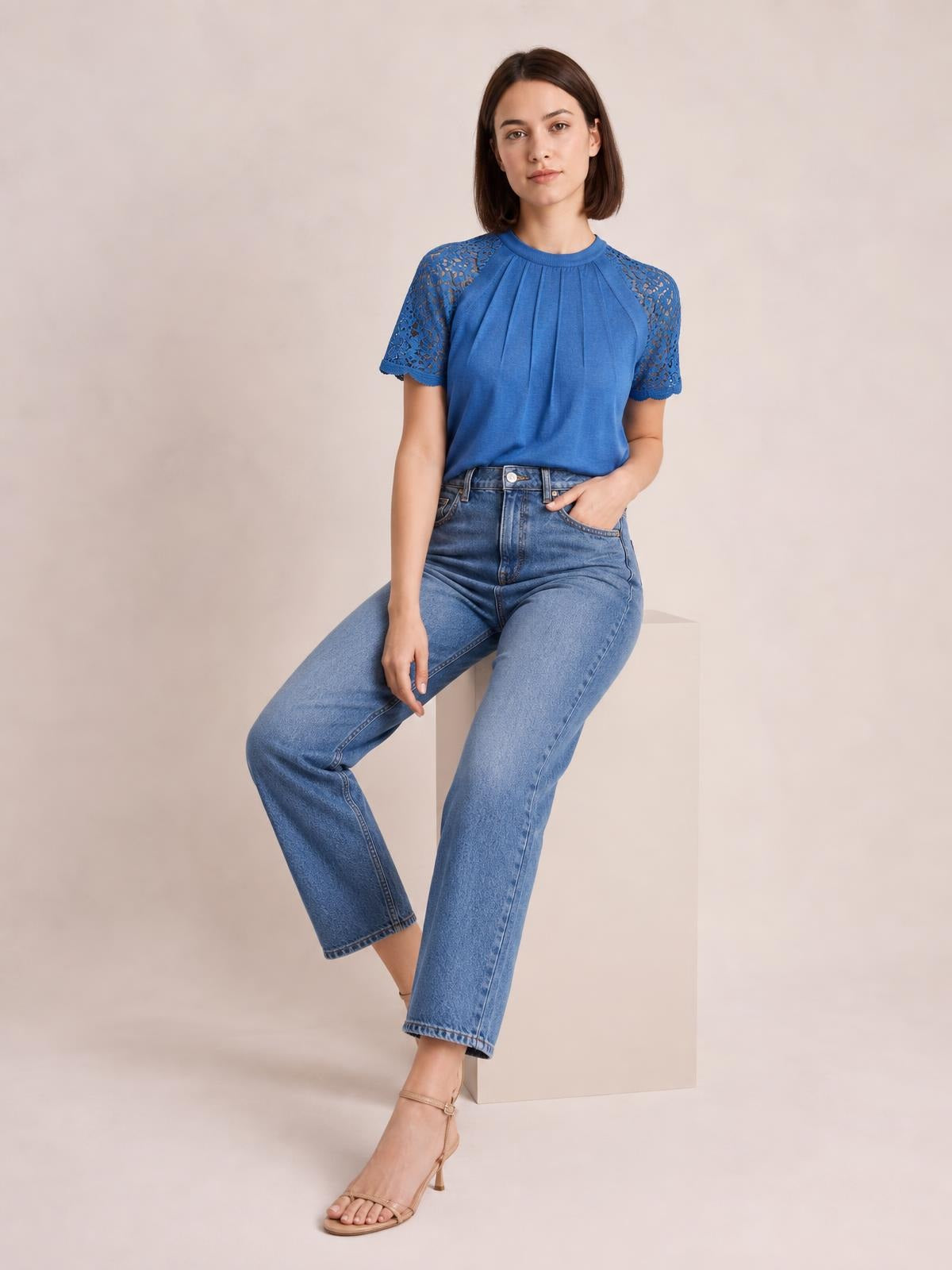 Model seated in a blue round neck short sleeve T-shirt with lace shoulder and sleeve details, pleated front, and a relaxed silhouette.