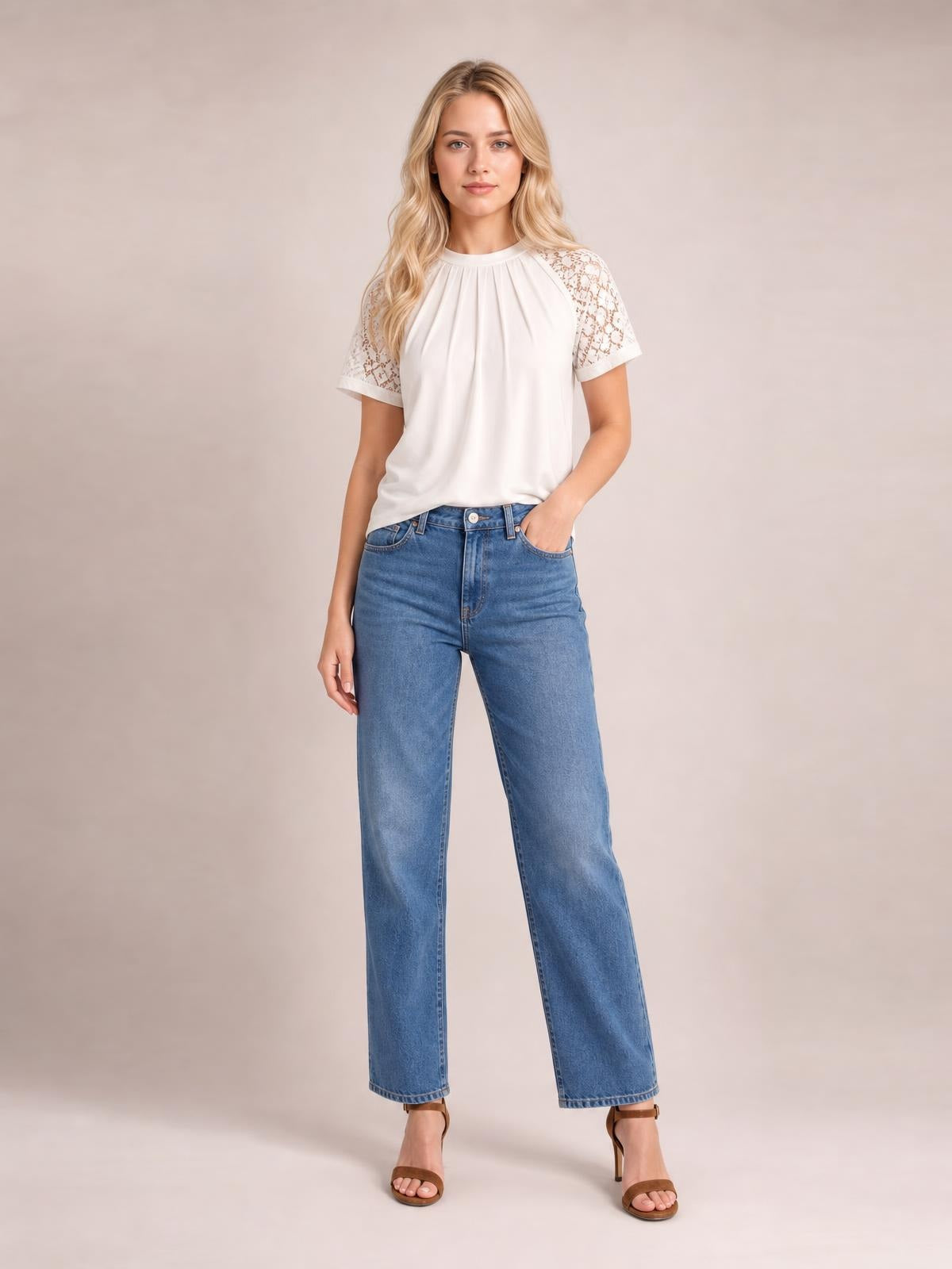 Model poses in a white round neck short sleeve T-shirt with lace shoulder panels, pleated front, and a relaxed silhouette tucked into blue jeans.