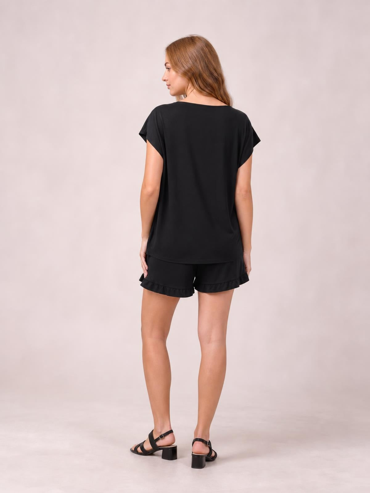 Model stands turned away in a black round-neck short-sleeve t-shirt and drawstring shorts lounge set with a relaxed silhouette and frilled hems.