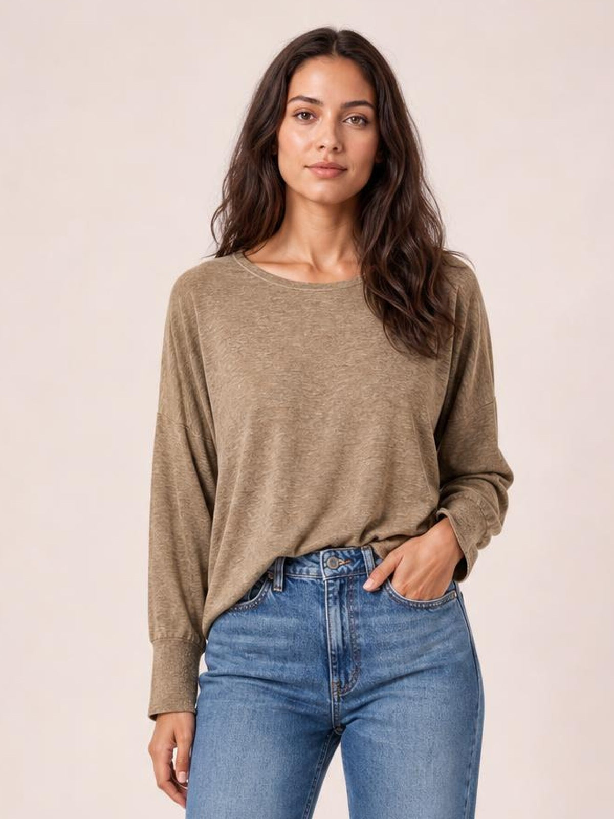 A model wears a taupe round-neck T-shirt with a relaxed silhouette, long lantern sleeves, and fitted cuffs, posing with one hand in her jeans pocket.