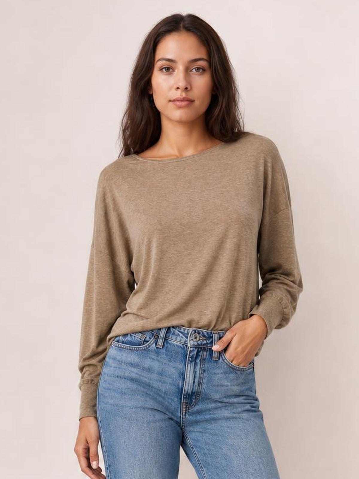 Model wearing a taupe round-neck T-shirt with relaxed long sleeves, a softly loose silhouette, and fitted cuffs, standing with one hand in her jeans pocket.