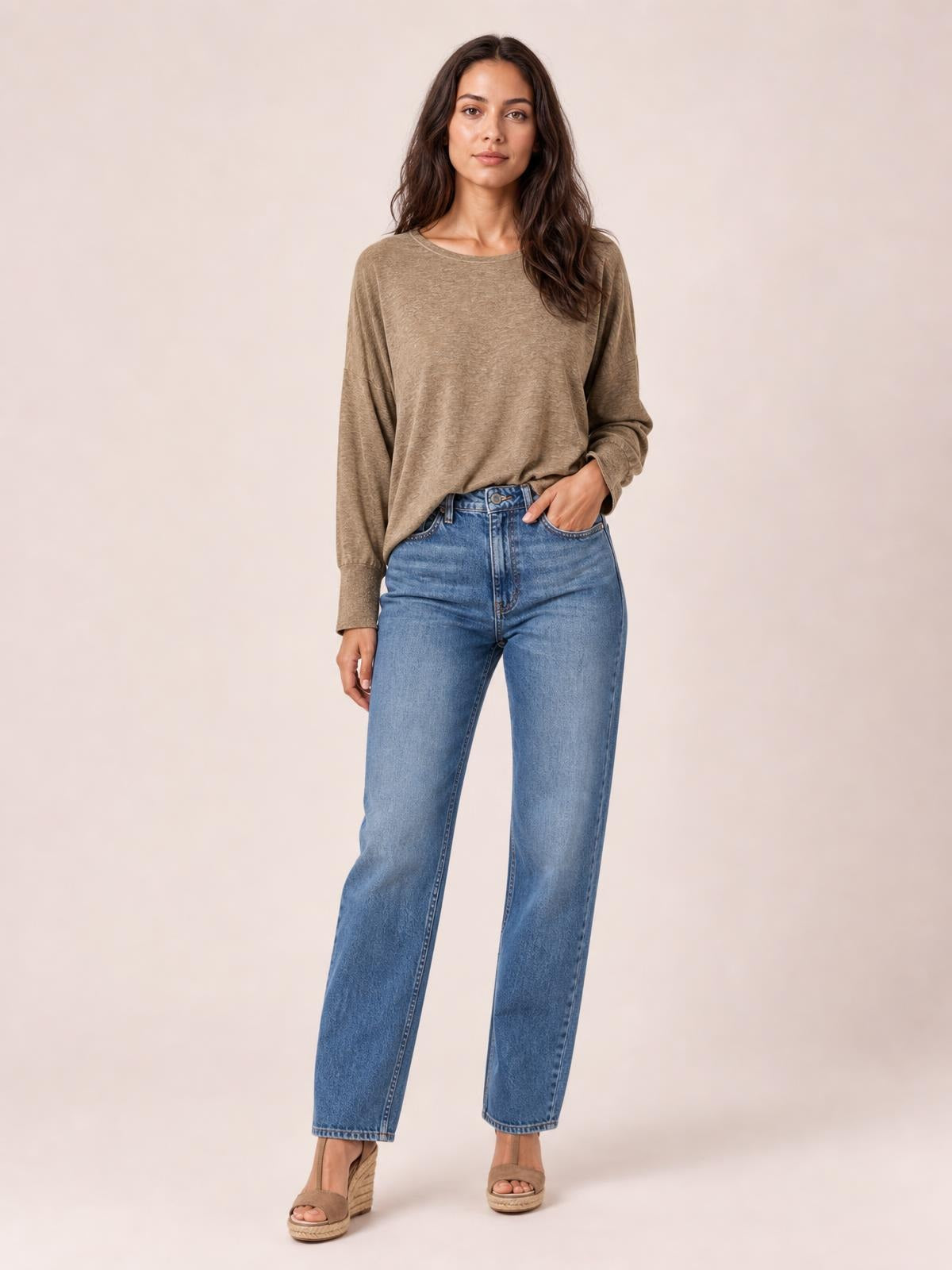 A model wears a taupe round neck smocked T-shirt with relaxed lantern sleeves and fitted cuffs, standing casually with one hand in her jeans pocket.