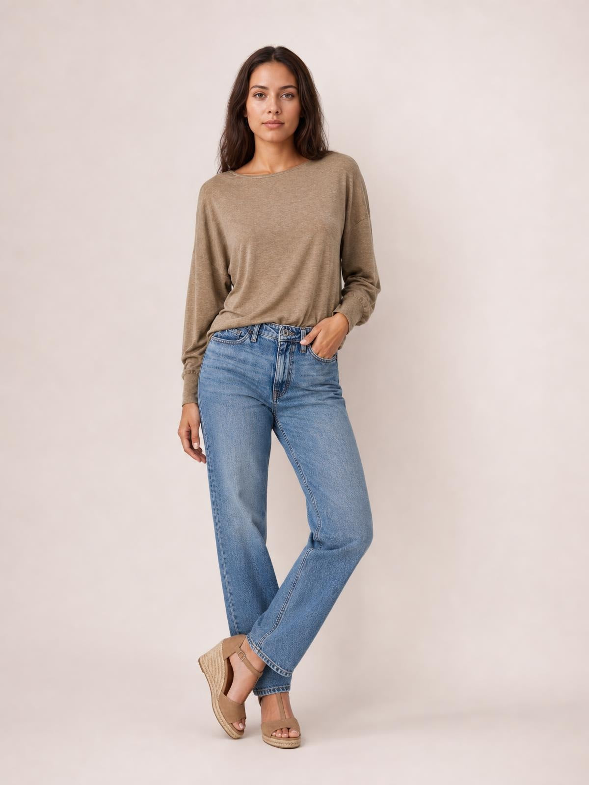 A model wears a taupe round-neck t-shirt with relaxed lantern sleeves and gathered cuffs, styled in a loose silhouette with one hand in her pocket.