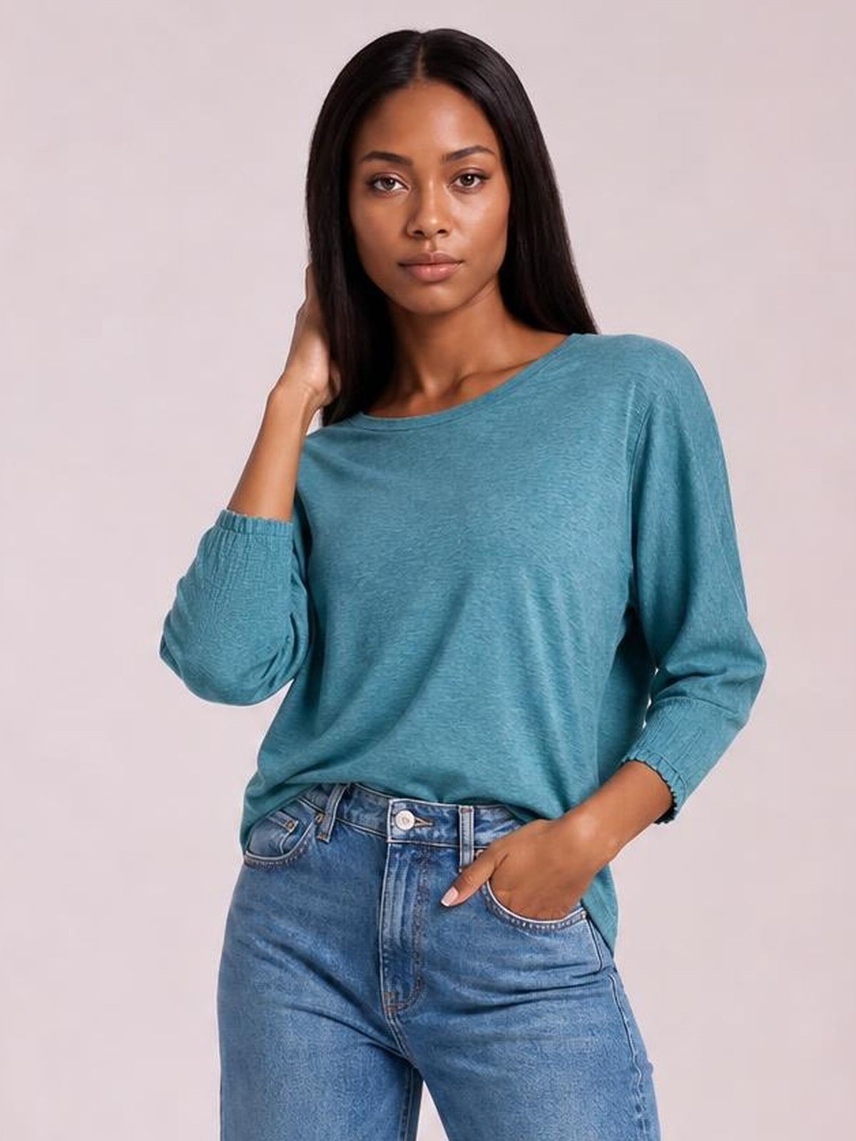 Model wears a teal round-neck T-shirt with a relaxed silhouette, long lantern sleeves, and smocked cuffs, posing with one hand in her jeans pocket.
