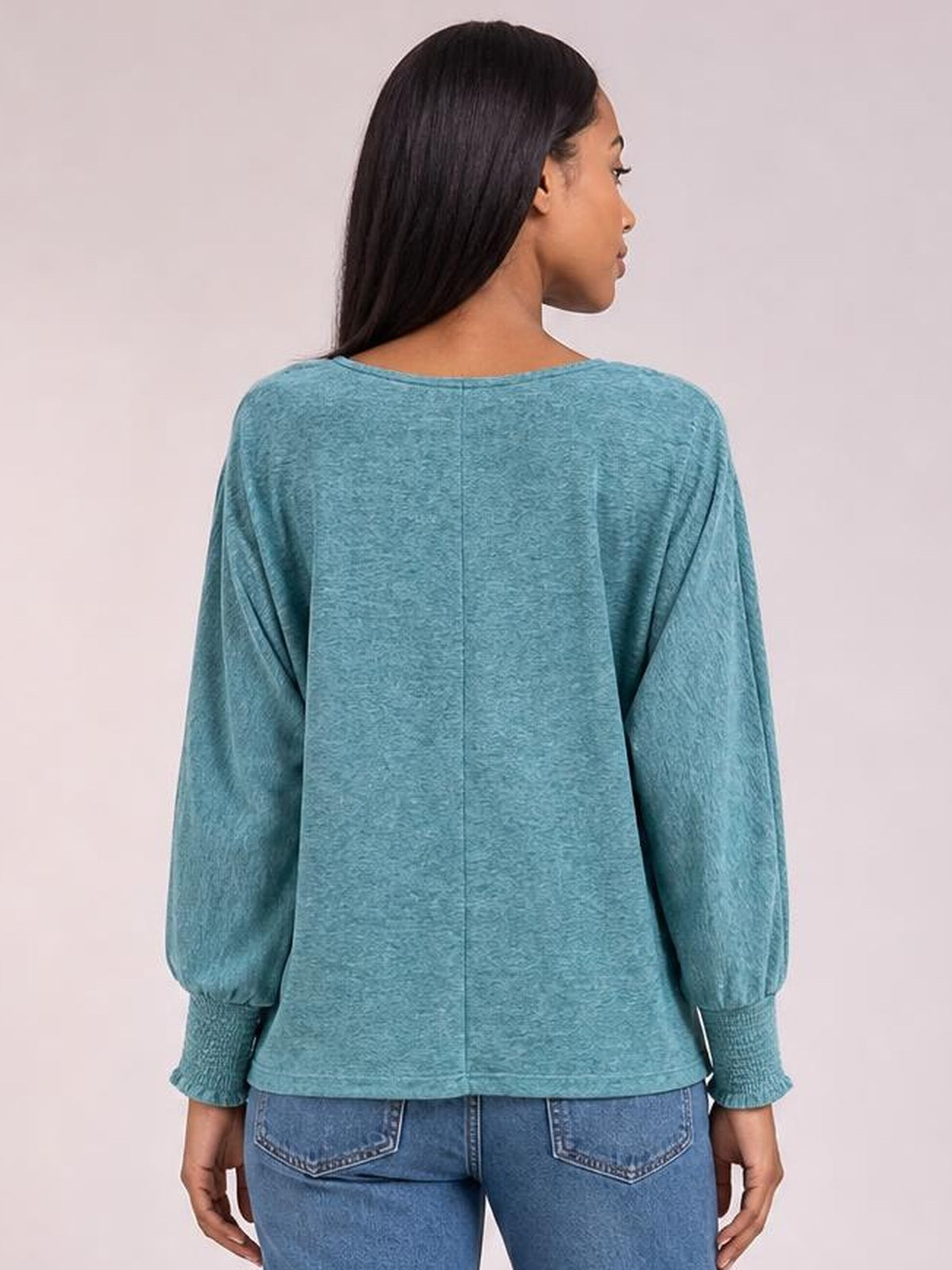 A model stands facing away in a teal round-neck T-shirt with a relaxed silhouette, lantern sleeves, smocked cuffs, and a straight hem, turning her head to the side.