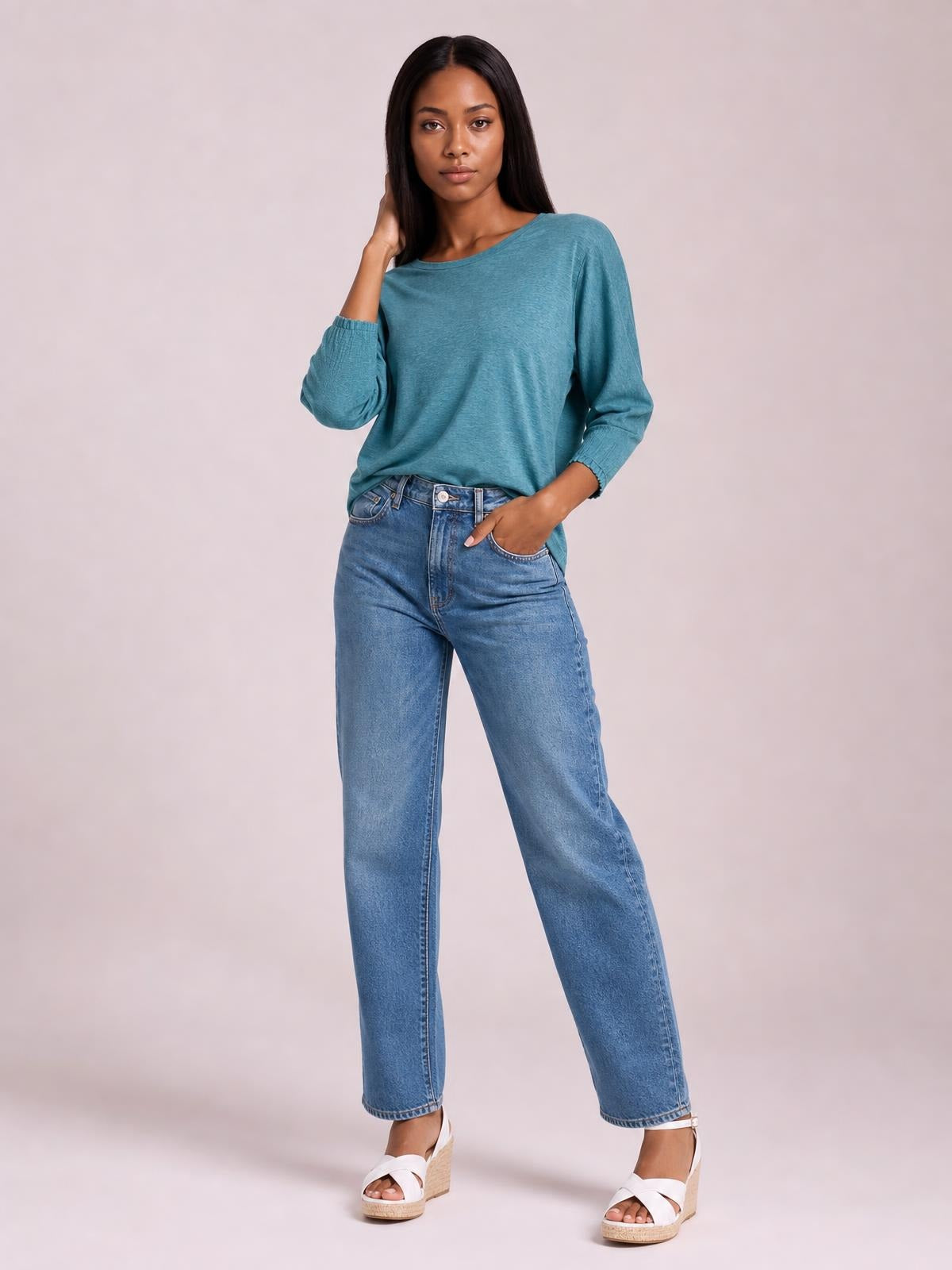 A model poses in a teal round-neck T-shirt with relaxed lantern sleeves, smocked cuffs, and a softly draped silhouette, touching her hair.