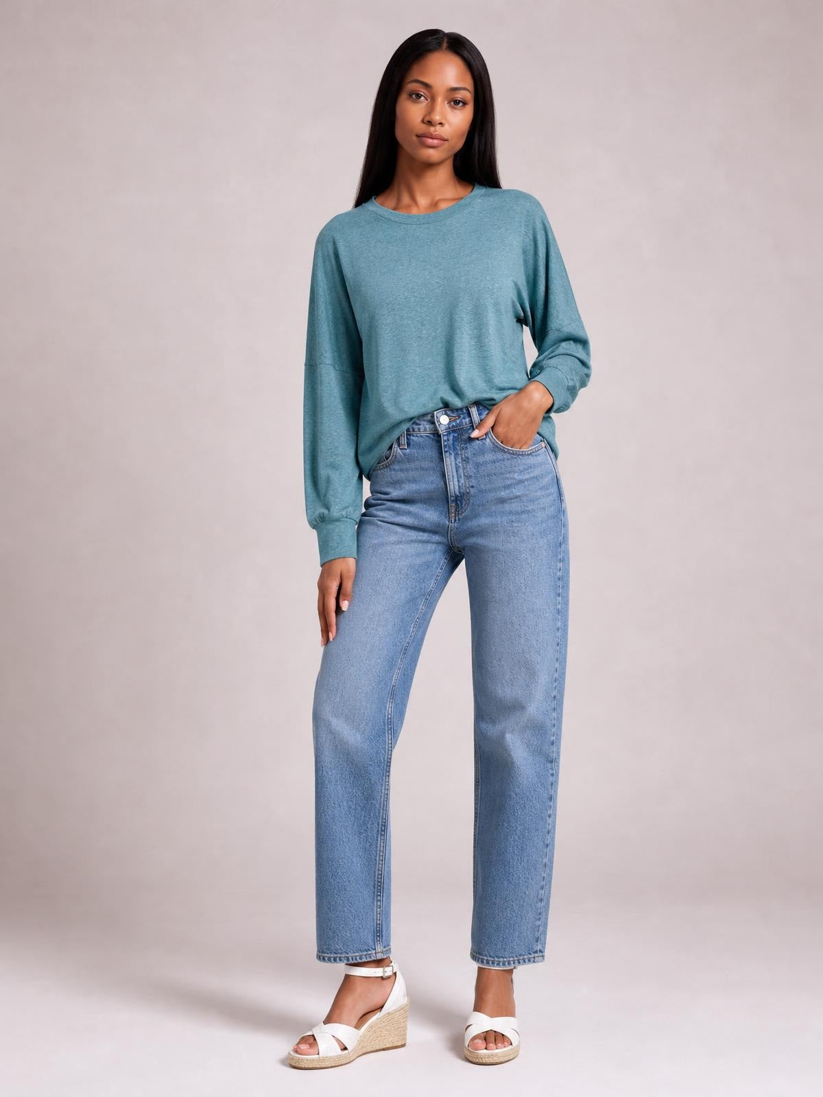 A model poses in a teal round-neck T-shirt with relaxed lantern sleeves, cuffed construction, a loose silhouette, and one hand tucked into her jeans pocket.