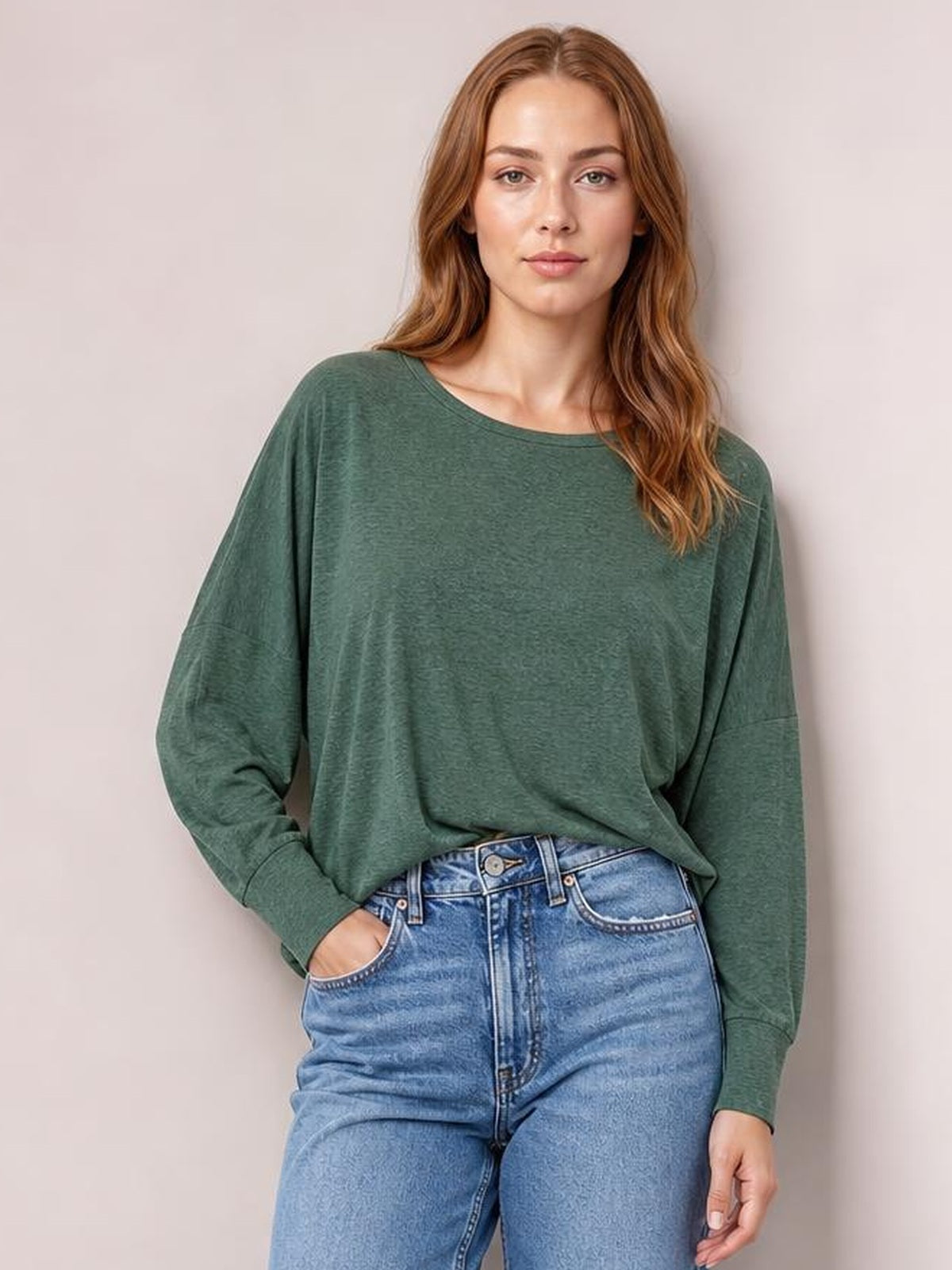 A model wears a sage green round neck smocked T-shirt with relaxed lantern sleeves and ribbed cuffs, standing front-facing with one hand in her jeans pocket.