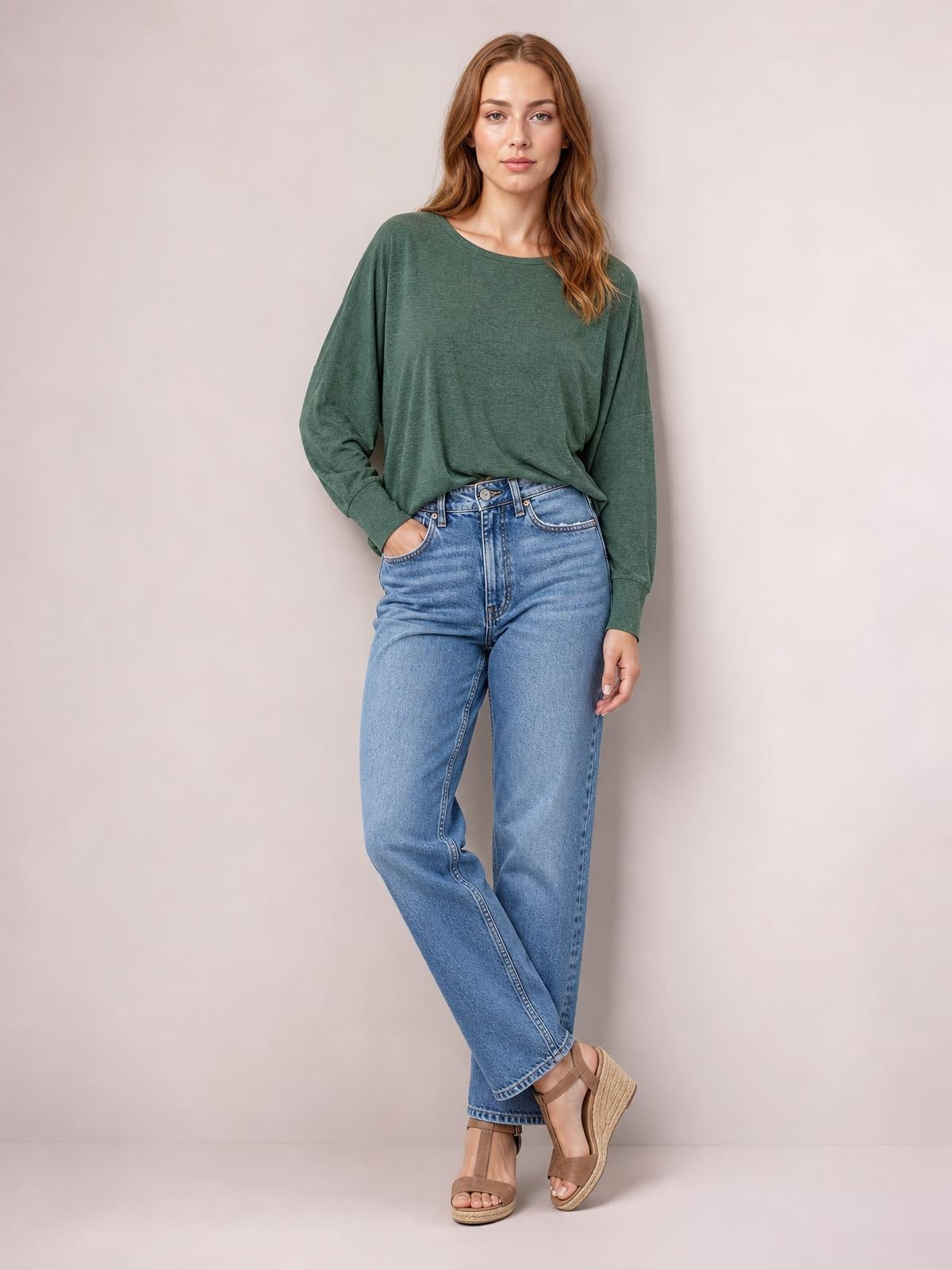 Model wears a forest-green round-neck T-shirt with relaxed long sleeves and a loose silhouette, posing with one hand in her pocket and crossed legs.
