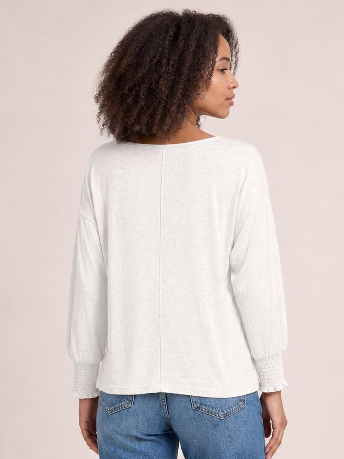 A model stands facing away in a white round-neck T-shirt with relaxed styling, long lantern sleeves, smocked cuffs, and a softly loose silhouette.