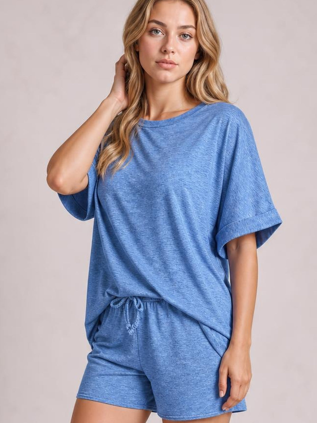A model poses with one hand by her hair in a blue round neck T-shirt and drawstring shorts set, featuring a relaxed oversized top and casual shorts.