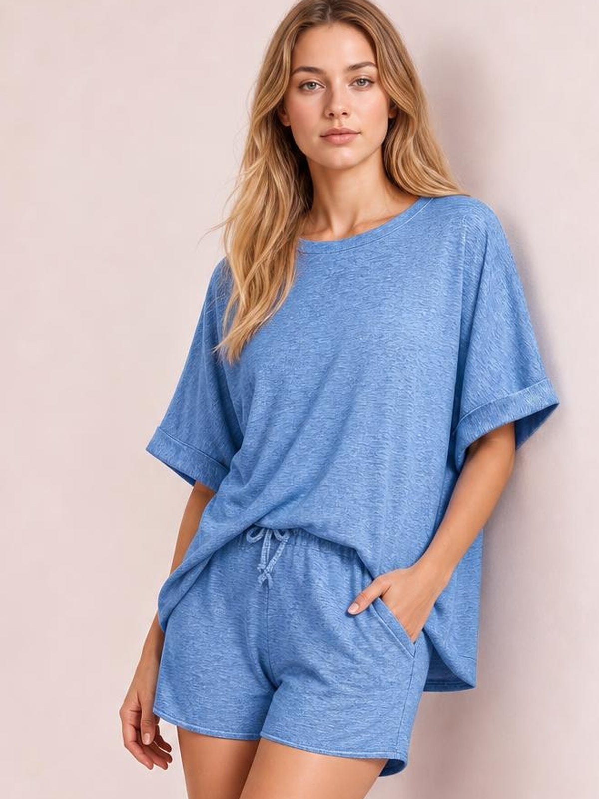 Model poses in a relaxed blue round neck T-shirt and drawstring shorts set, featuring an oversized top, rolled sleeves, elastic waist, and casual loose silhouette.