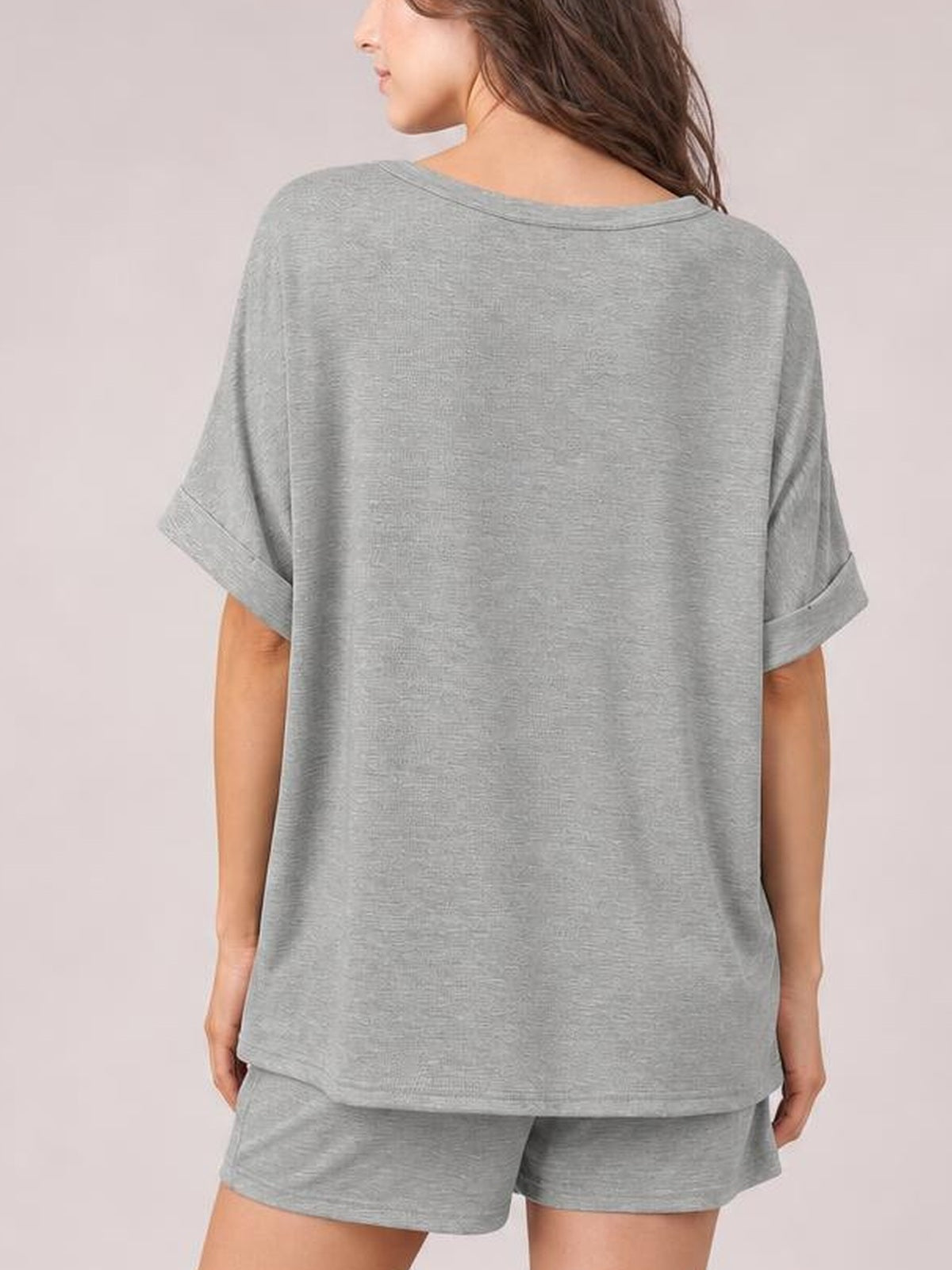 Model shown from behind wears a relaxed gray round neck T-shirt with rolled short sleeves and matching drawstring shorts in a casual set silhouette.