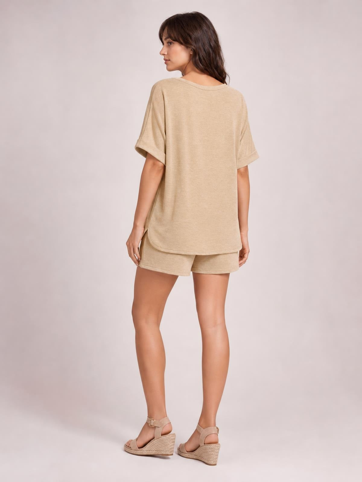 Model stands turned away in a beige round neck T-shirt and drawstring shorts set, featuring a relaxed silhouette, rolled short sleeves, and curved hem.