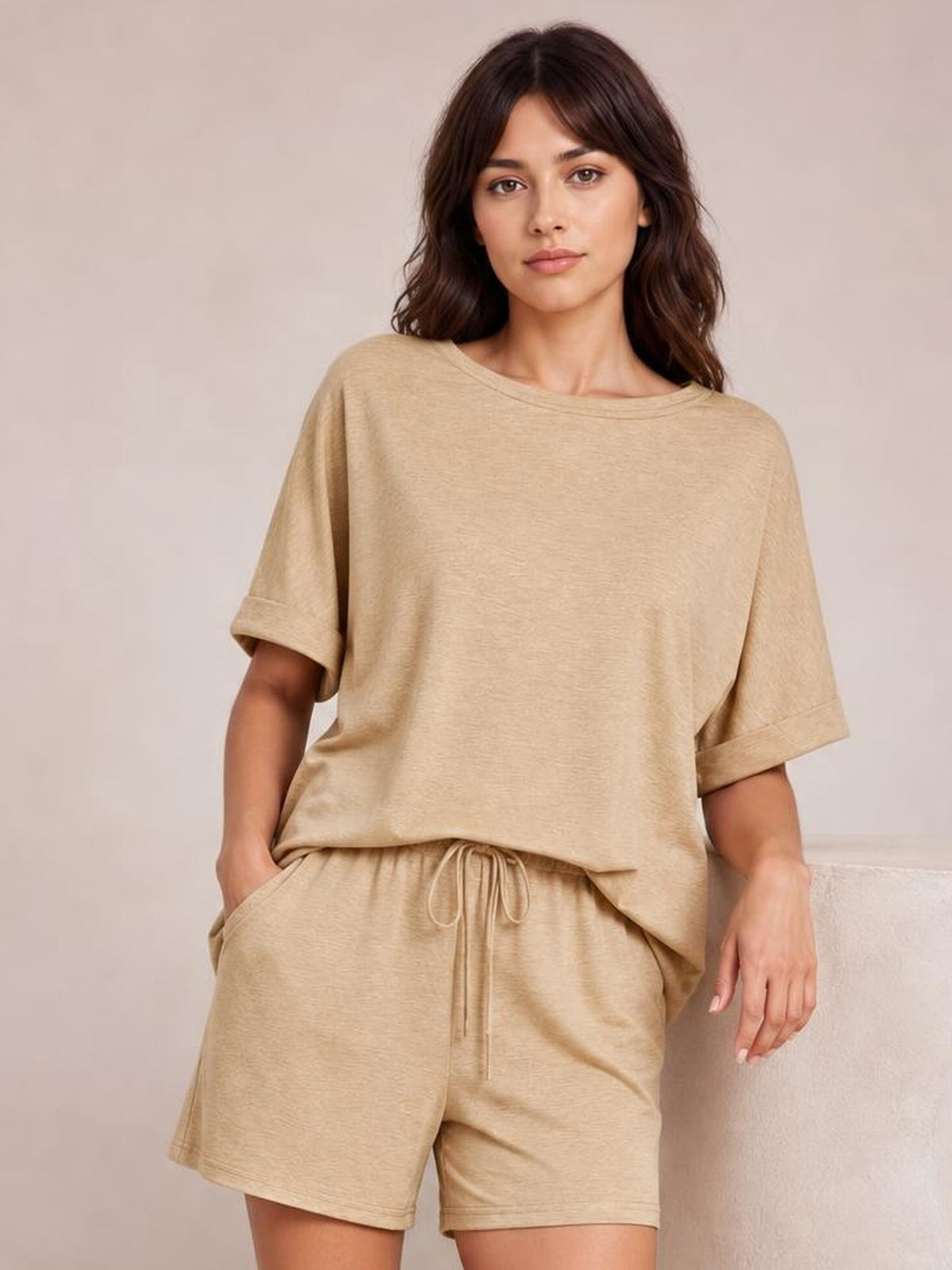 Model wears a beige round neck T-shirt and drawstring shorts set with a relaxed oversized silhouette, rolled sleeves, side pockets, and one hand tucked in a pocket.