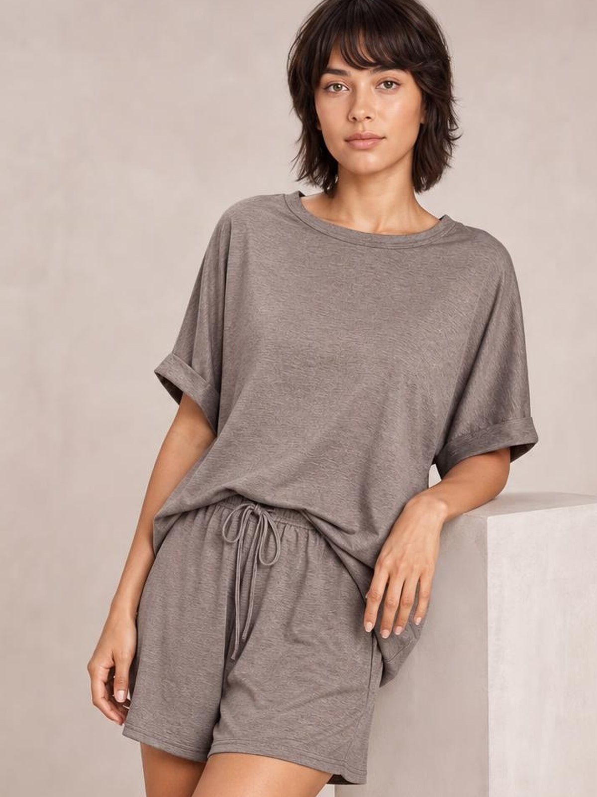 A model poses in a taupe round neck T-shirt and drawstring shorts set with an oversized silhouette, rolled short sleeves, and relaxed shorts.