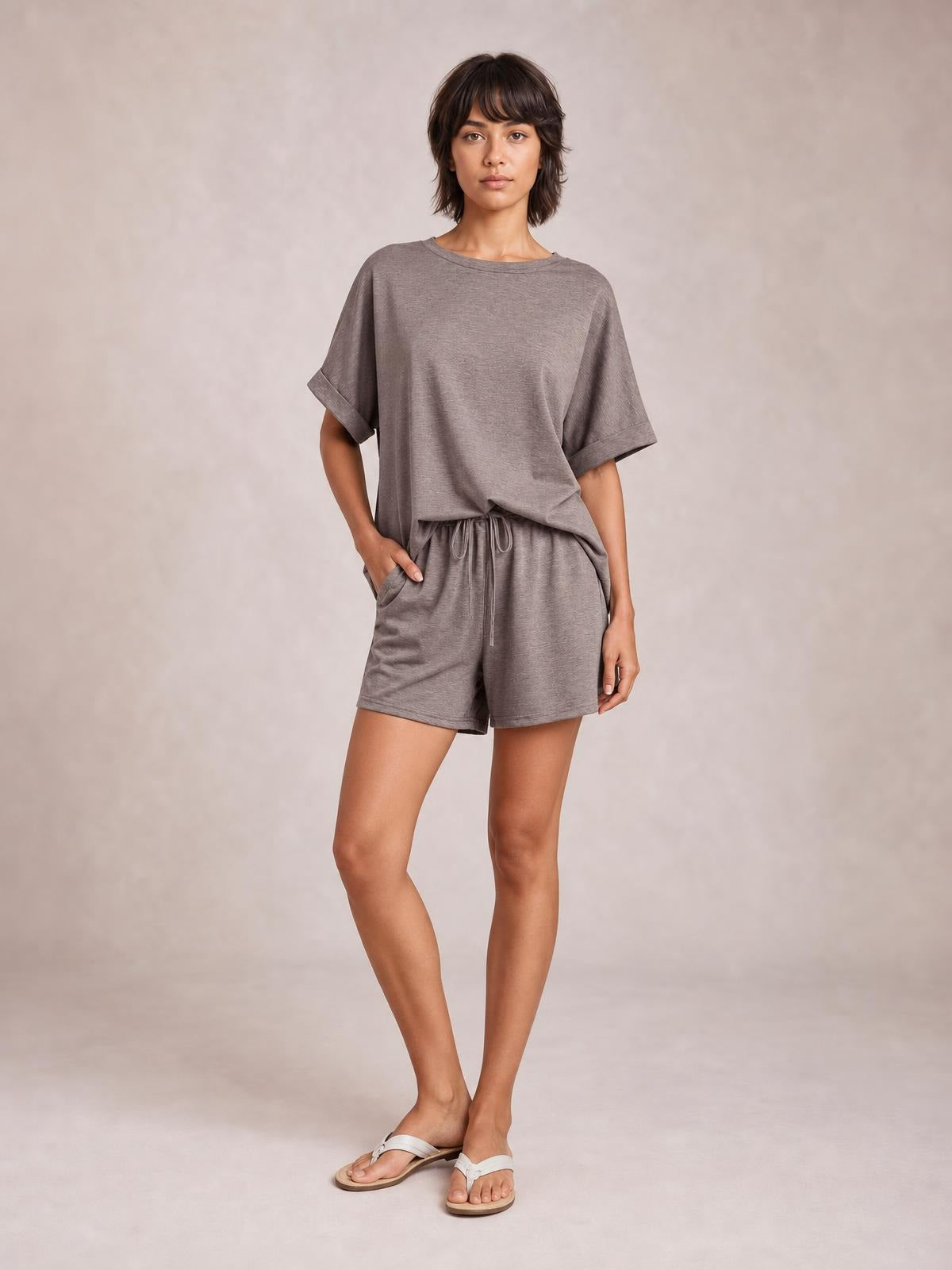 Model wears a taupe round neck T-shirt and drawstring shorts set with an oversized top, rolled sleeves, relaxed shorts, and one hand in a pocket.
