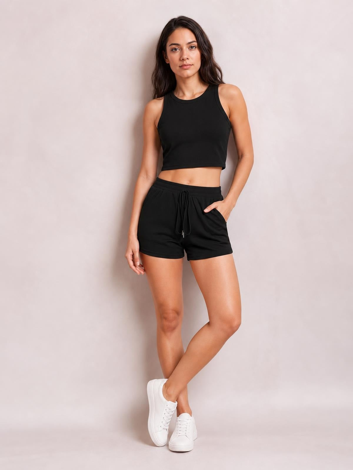 A woman models a black round neck tank and drawstring shorts set with a cropped silhouette, side pockets, and a relaxed crossed-leg pose.