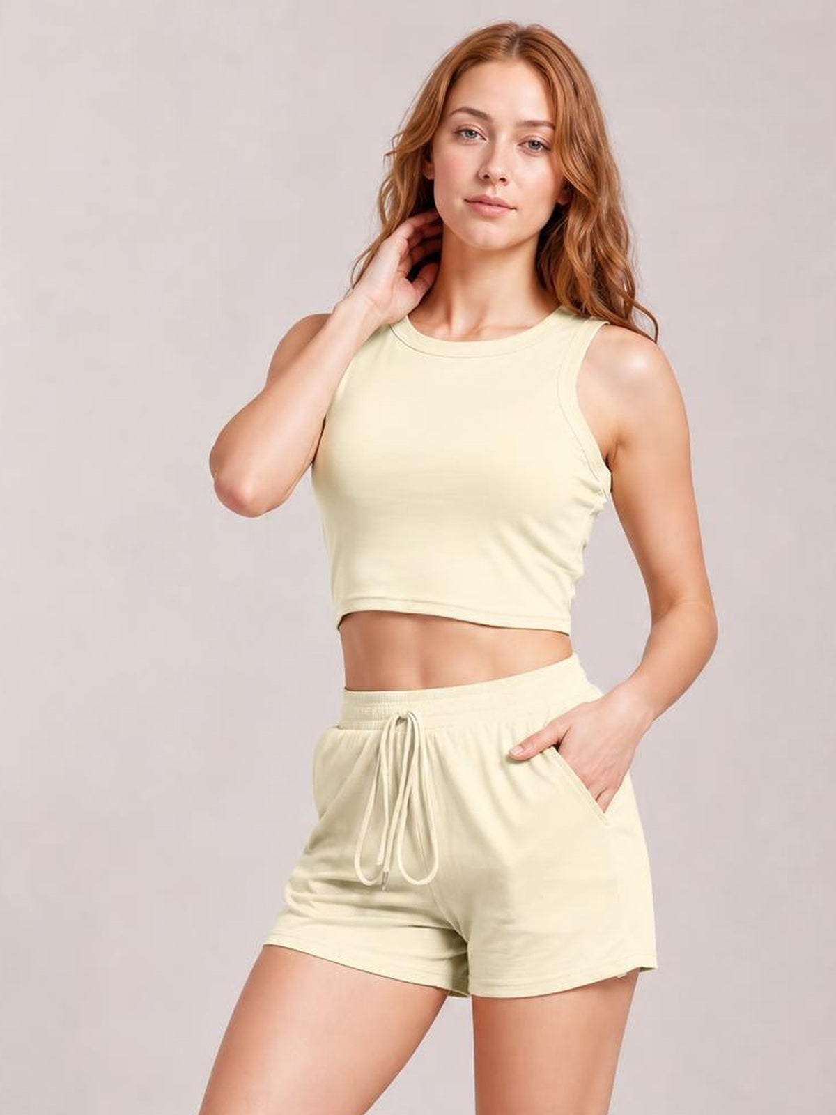 A model poses in a pale yellow round-neck tank and drawstring shorts set with a fitted cropped silhouette, side pockets, and one hand in a pocket.
